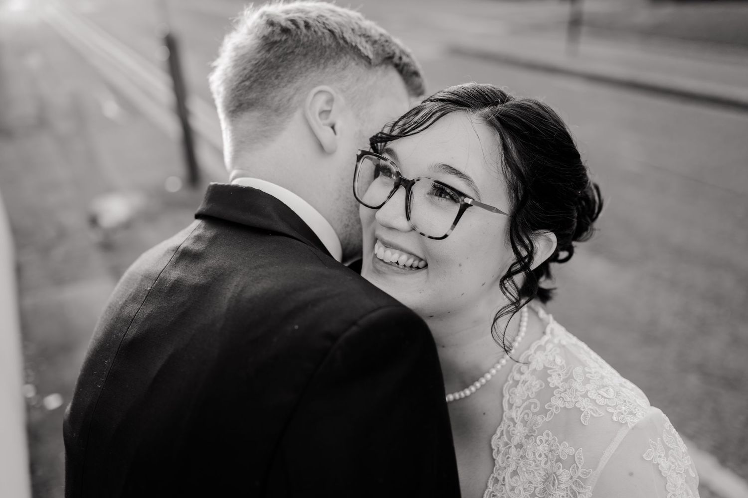 Black and white wedding photo of smiling bride in lace dress and glasses leaning close to groom in dark suit.