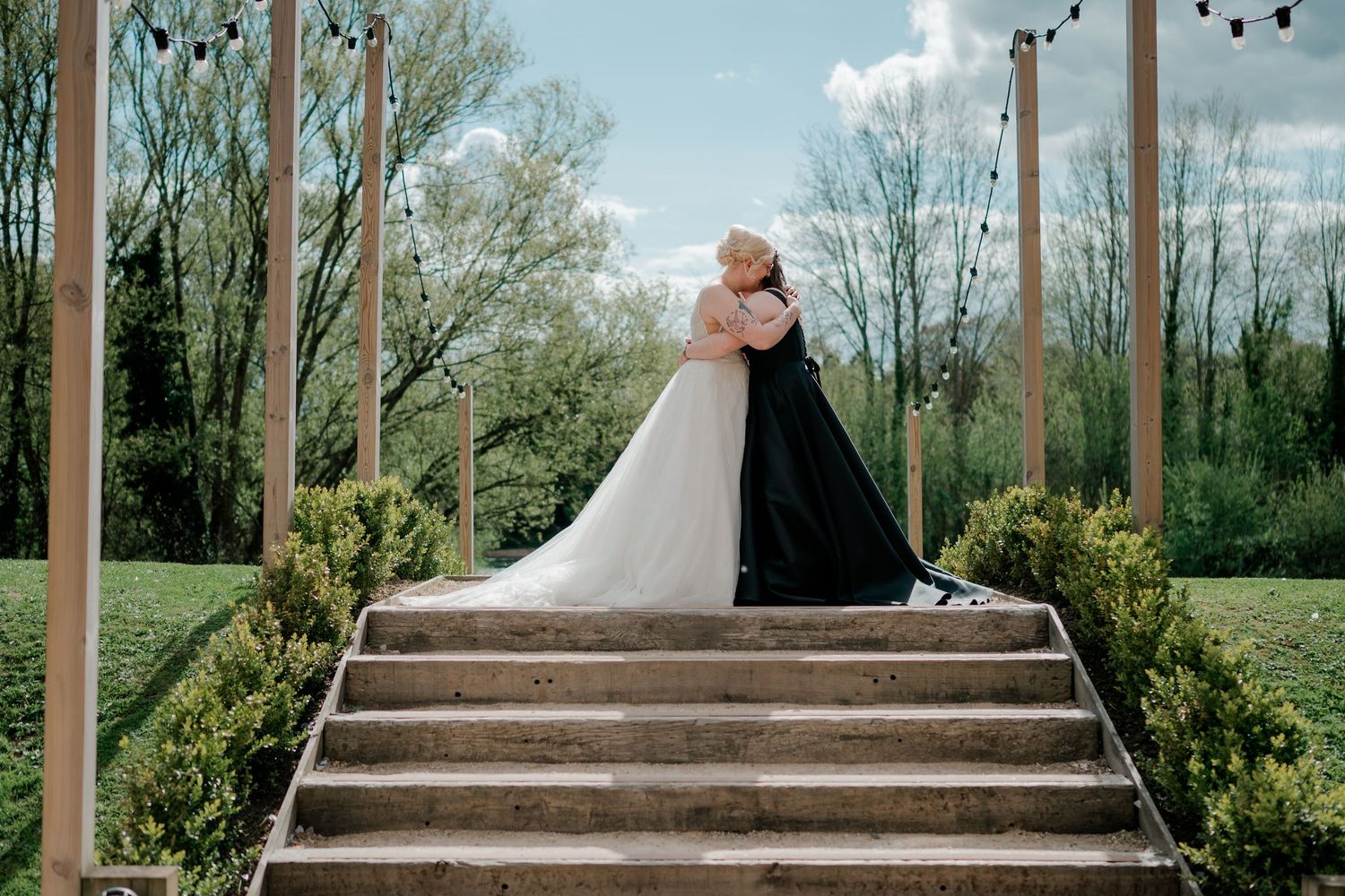 Bride and groom embracing at the top of wooden stairs surrounded by greenery and string lights at an outdoor wedding venue.