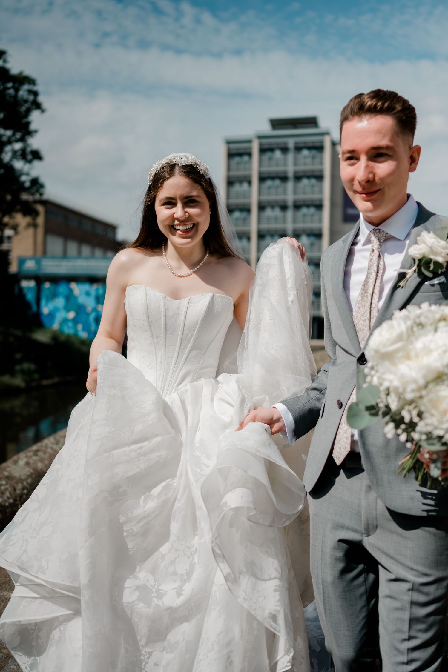 Smiling bride in strapless gown and groom in gray suit holding hands outdoors on their wedding day.