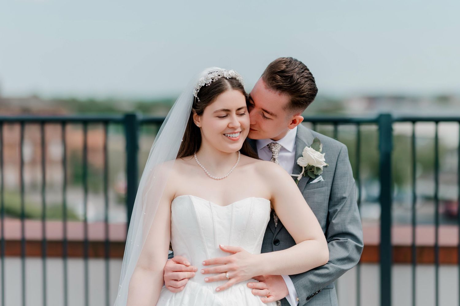 A groom in a gray suit kisses his bride in a white gown and veil on a rooftop at their wedding.