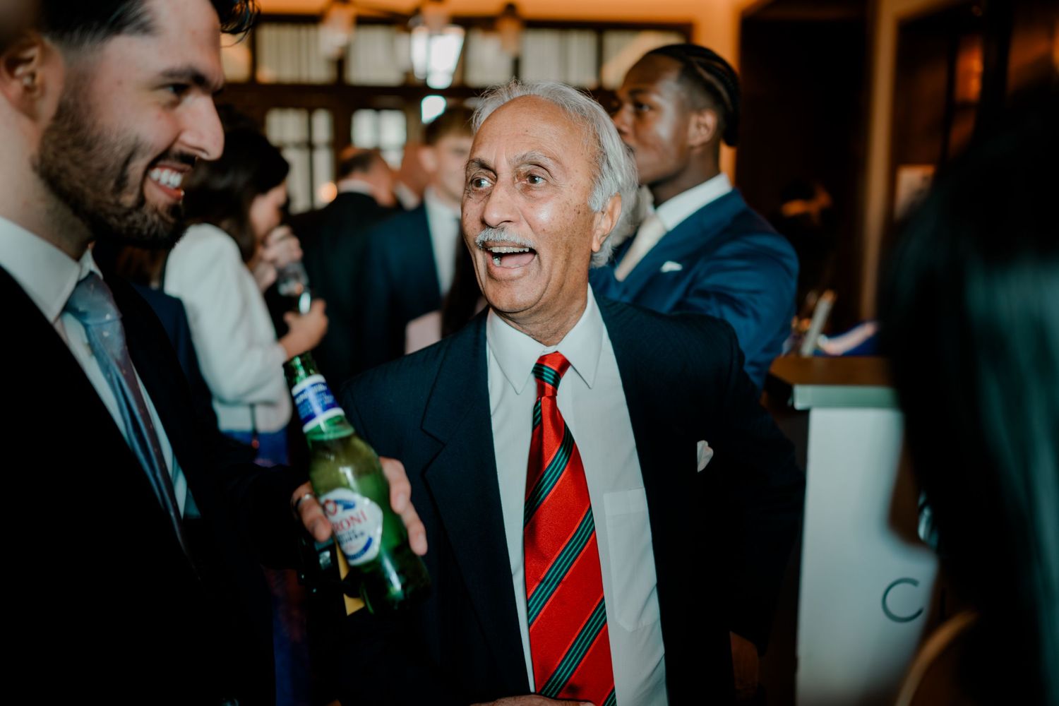 Elderly man in suit and striped tie laughing at a social gathering while holding a green beer bottle.