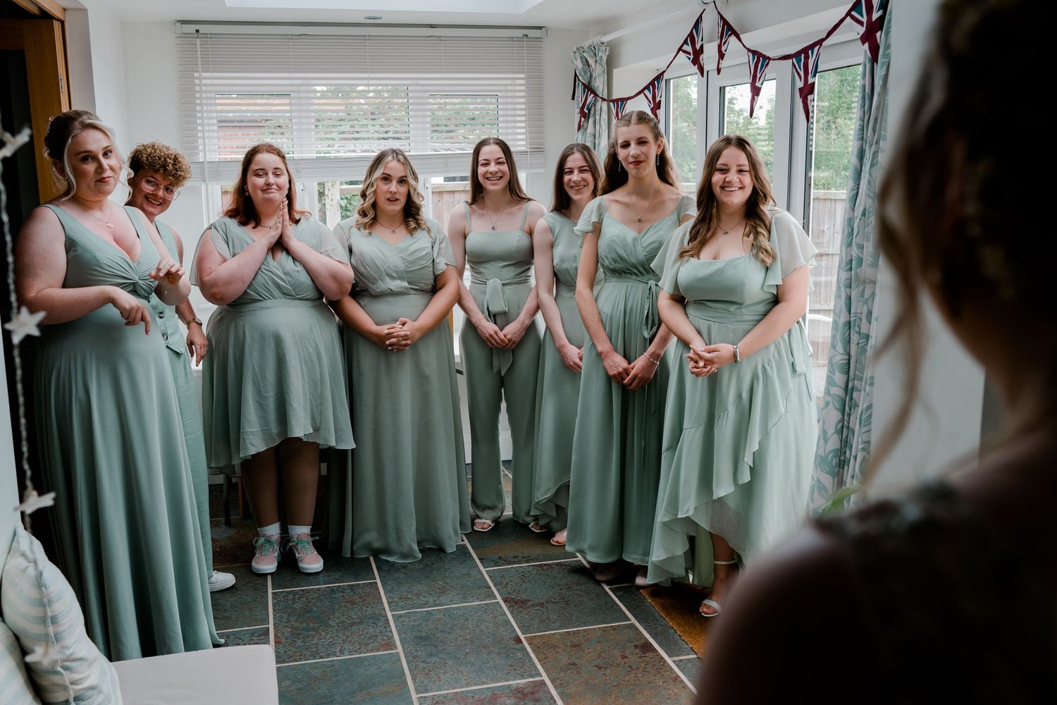 Six bridesmaids in matching sage green dresses smiling at the bride during wedding preparations.