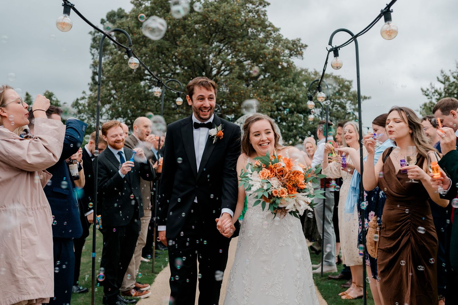 Wedding guests blow bubbles during an outdoor celebration as newlyweds walk through the joyful scene.