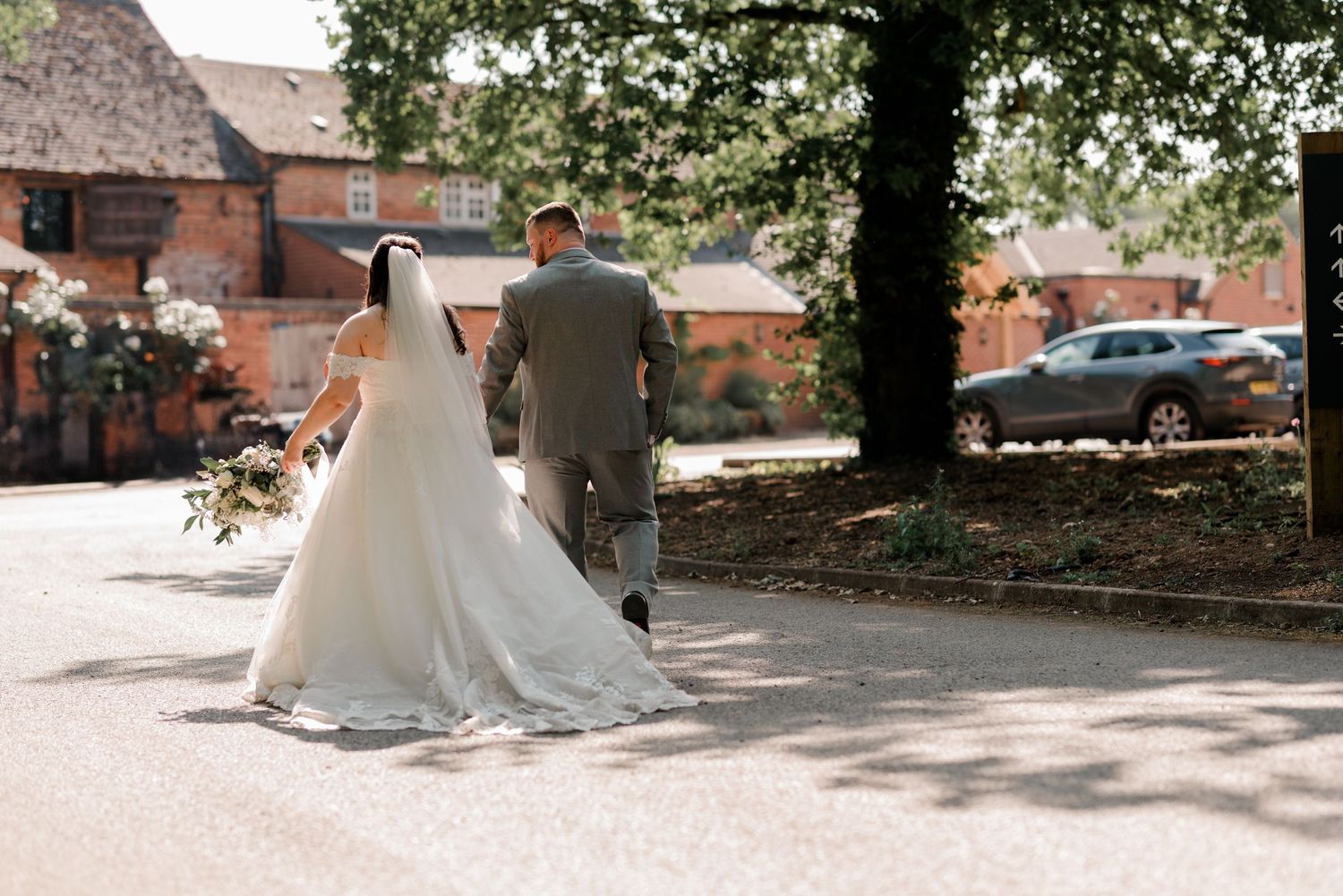 Newlyweds walking together on a sunny day with brick buildings and trees in the background.