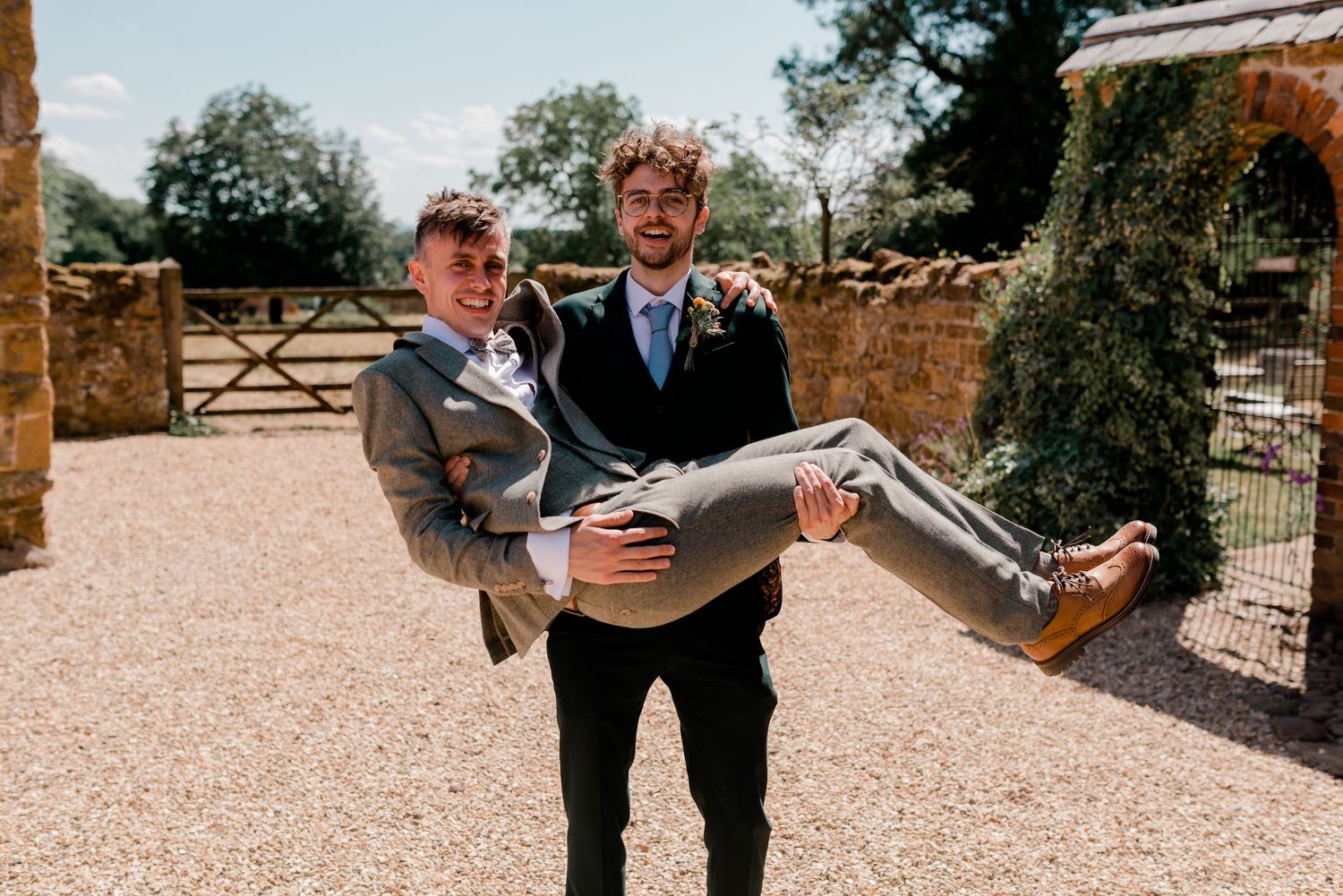 Wedding party members walk and mingle outside on a gravel path near rustic wooden gates.