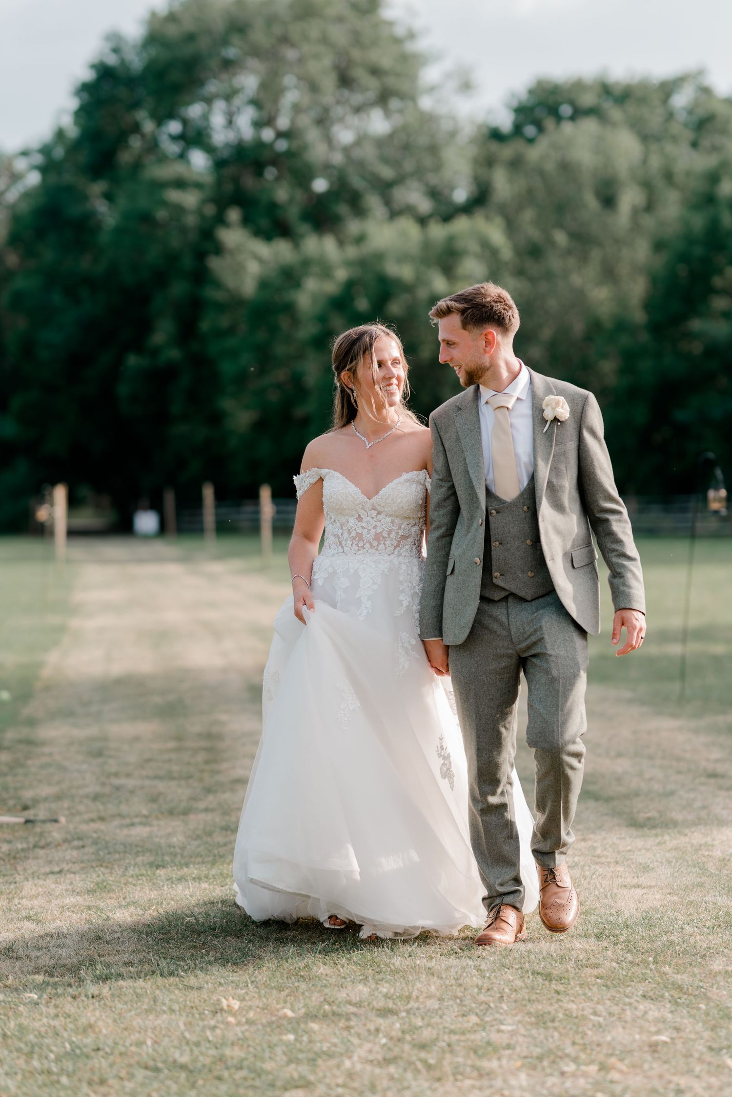 Smiling bride in off-shoulder lace gown walks with groom in grey suit through sunlit outdoor wedding venue.