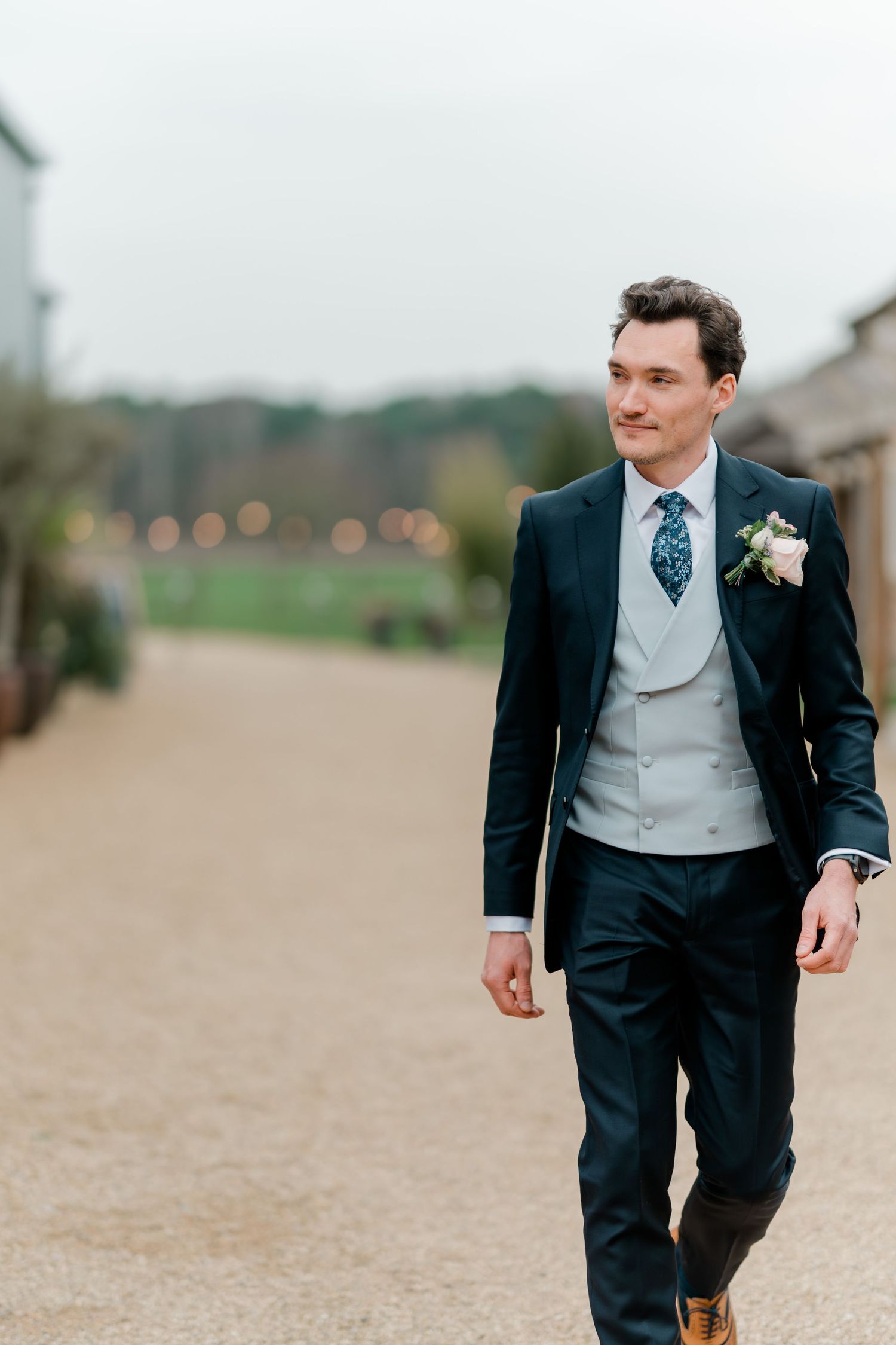 Dapper groom in navy suit with grey waistcoat and floral boutonniere walking outdoors at wedding venue.