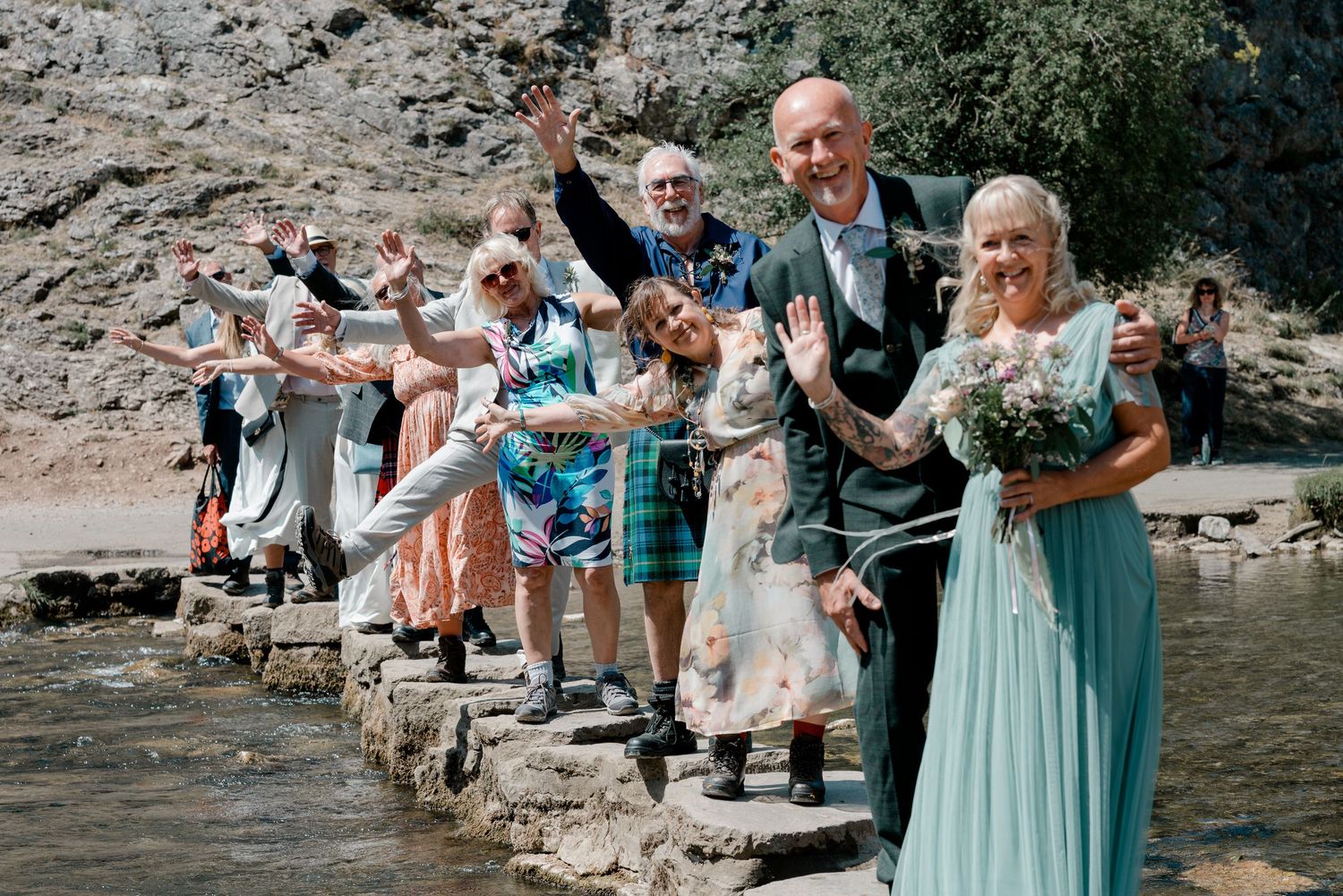 Wedding party posing on stepping stones by a river, bride in teal dress holding bouquet, guests cheering outdoors.