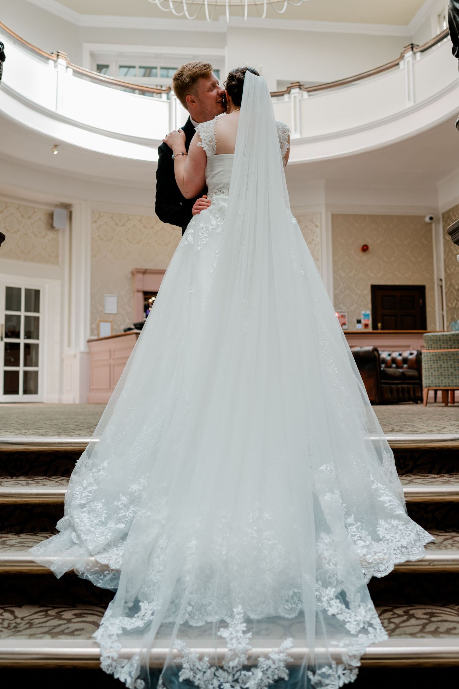 Bride in white lace ball gown with veil kisses groom in black suit on grand staircase in elegant venue.