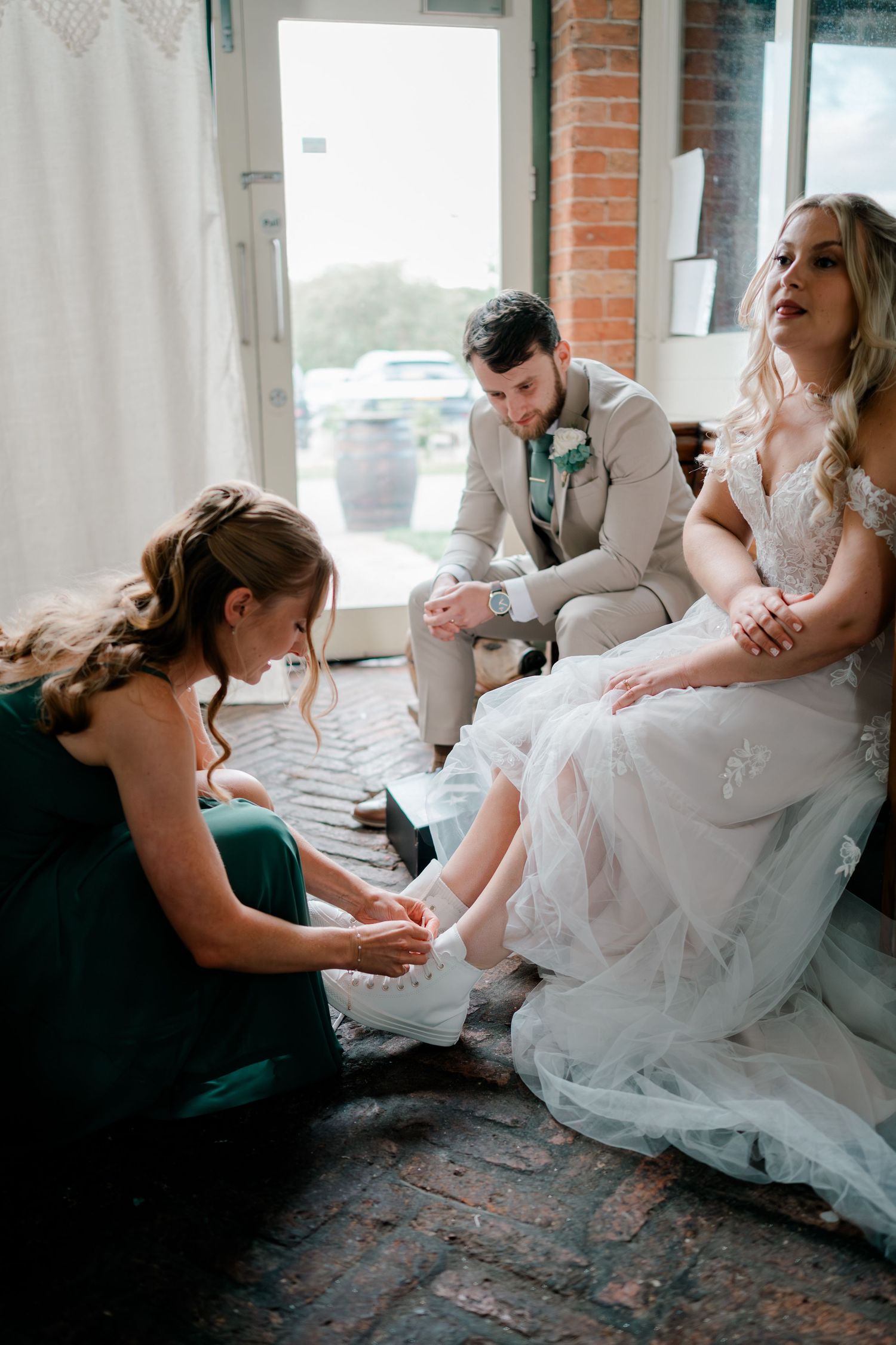 Bridesmaid in green dress helps bride with wedding shoes while groom watches in rustic brick venue.