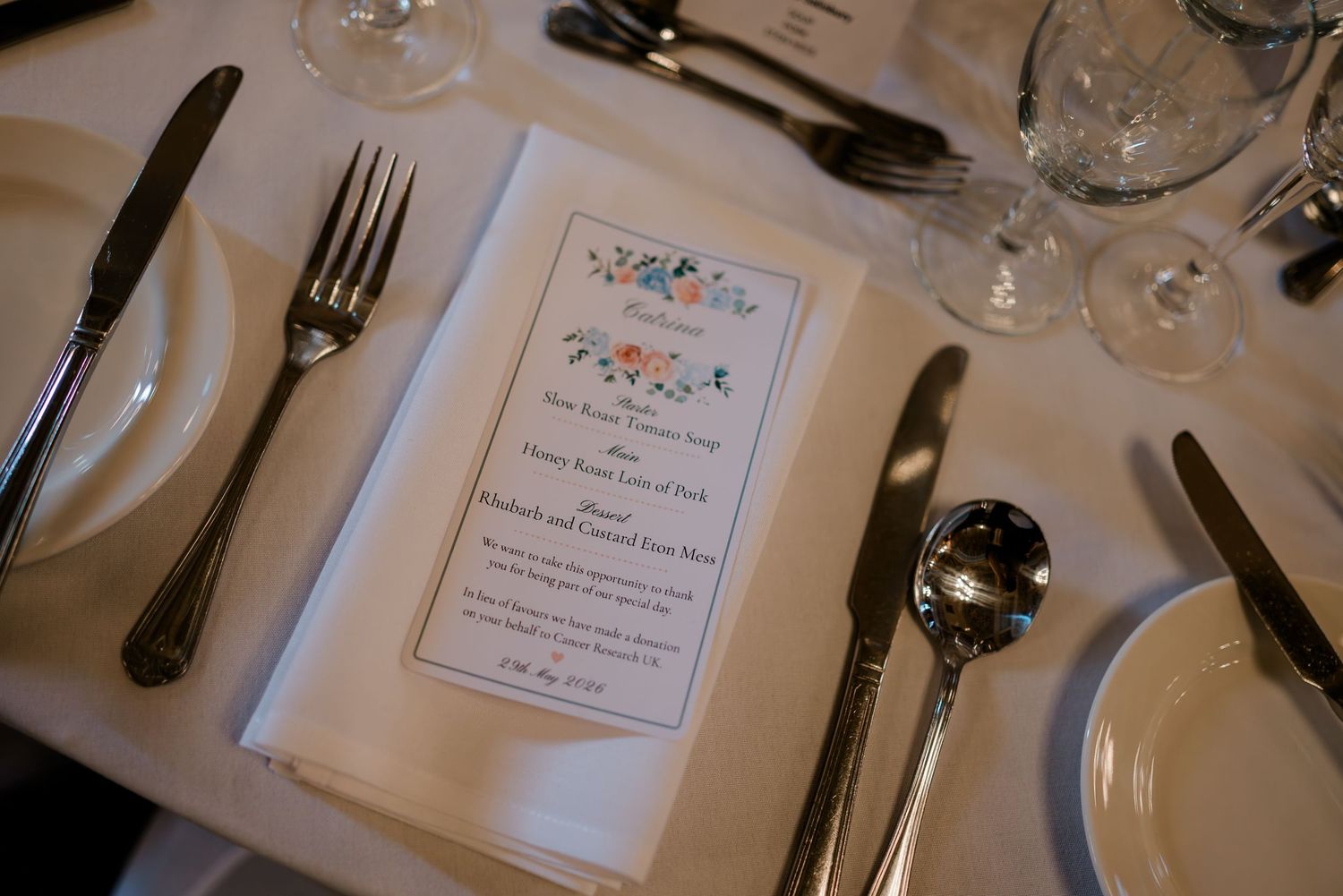 Elegant wedding reception place setting with floral menu card, silverware, and crystal glasses on white tablecloth.