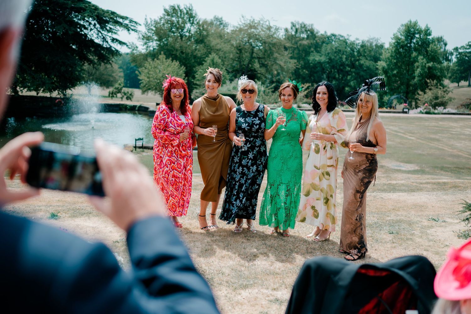 Six women in colorful summer dresses pose with drinks on a sunny lawn by a fountain while a guest photographs them at an outdoor wedding.