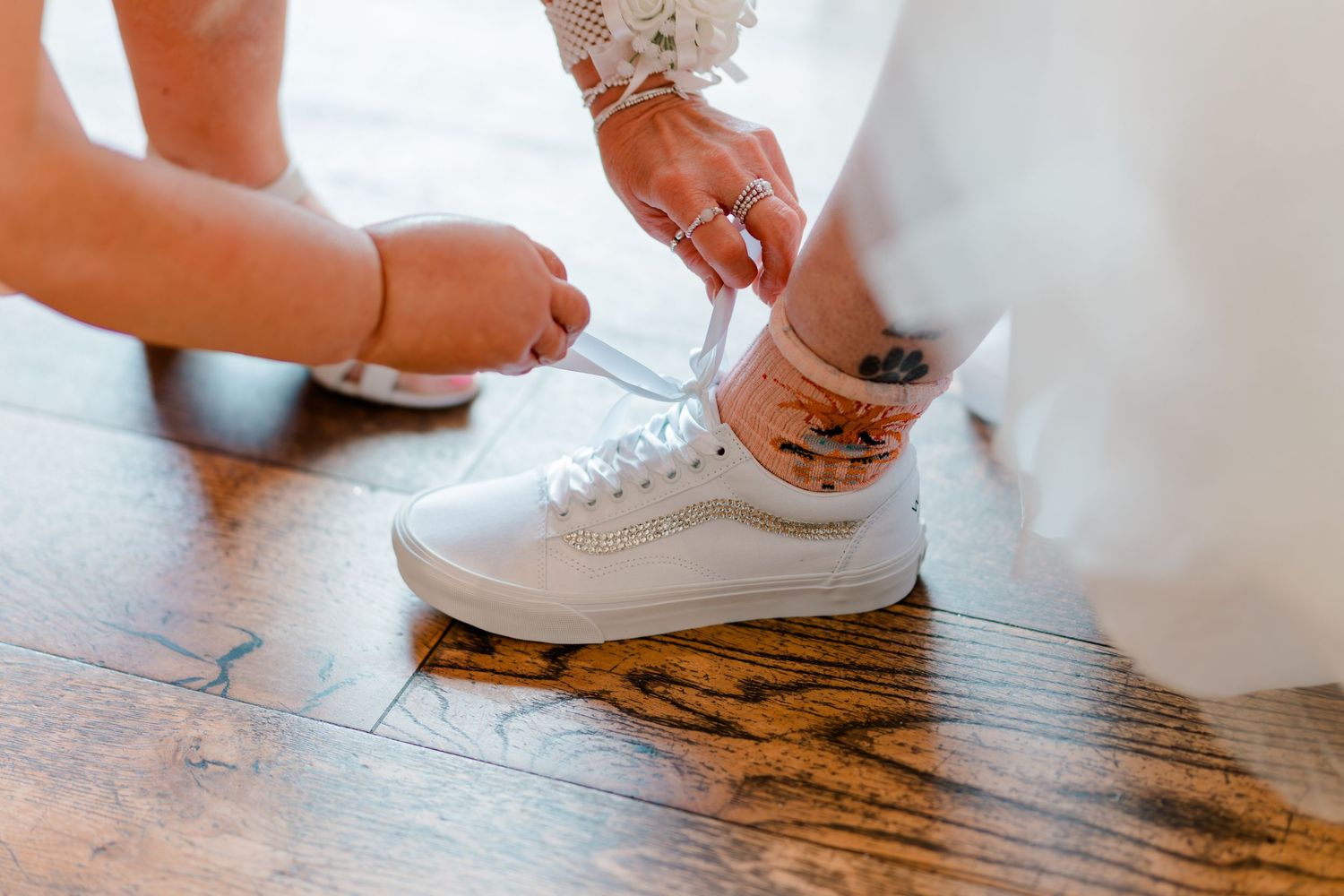 Bride tying white Vans sneakers on wood floor, showing ankle tattoo and bracelet, wearing wedding dress.