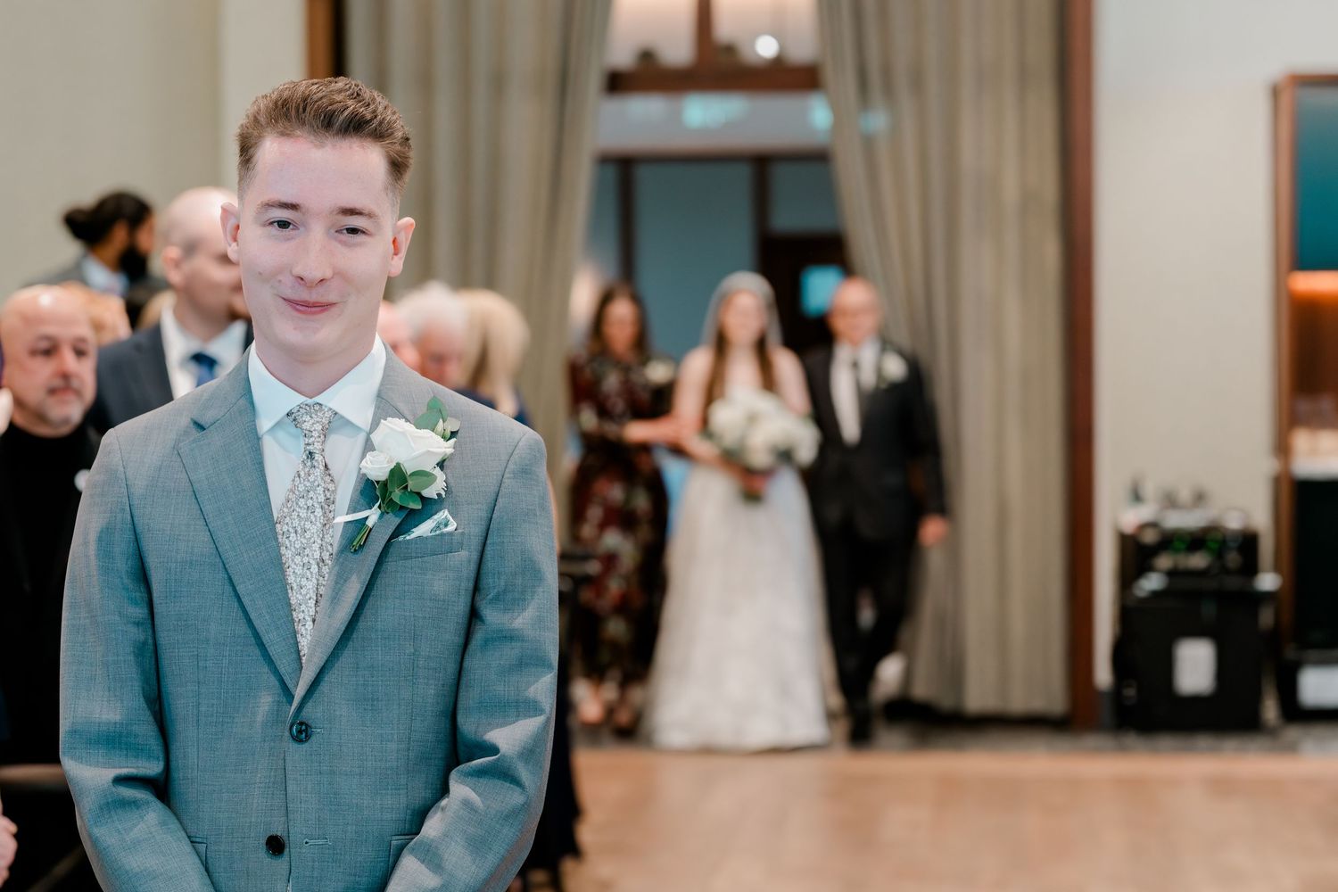 Groom in gray suit with boutonniere stands at altar as bride walks down aisle with escort at wedding ceremony.