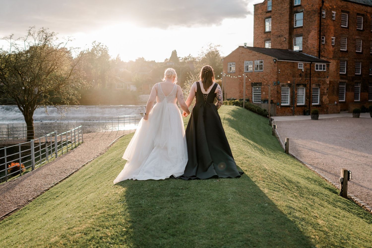 Two brides in white and black gowns walk hand-in-hand at golden hour beside a historic brick venue.