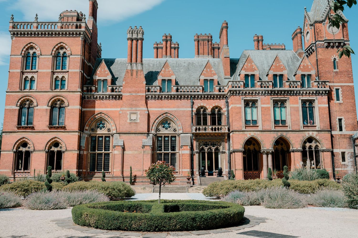 Grand Victorian red brick mansion with Gothic arched windows, manicured hedges, and circular garden feature.