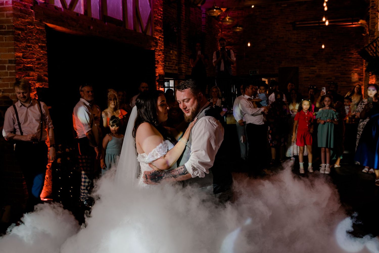 Dancing couple surrounded by atmospheric fog on a dance floor at a dimly lit wedding venue.