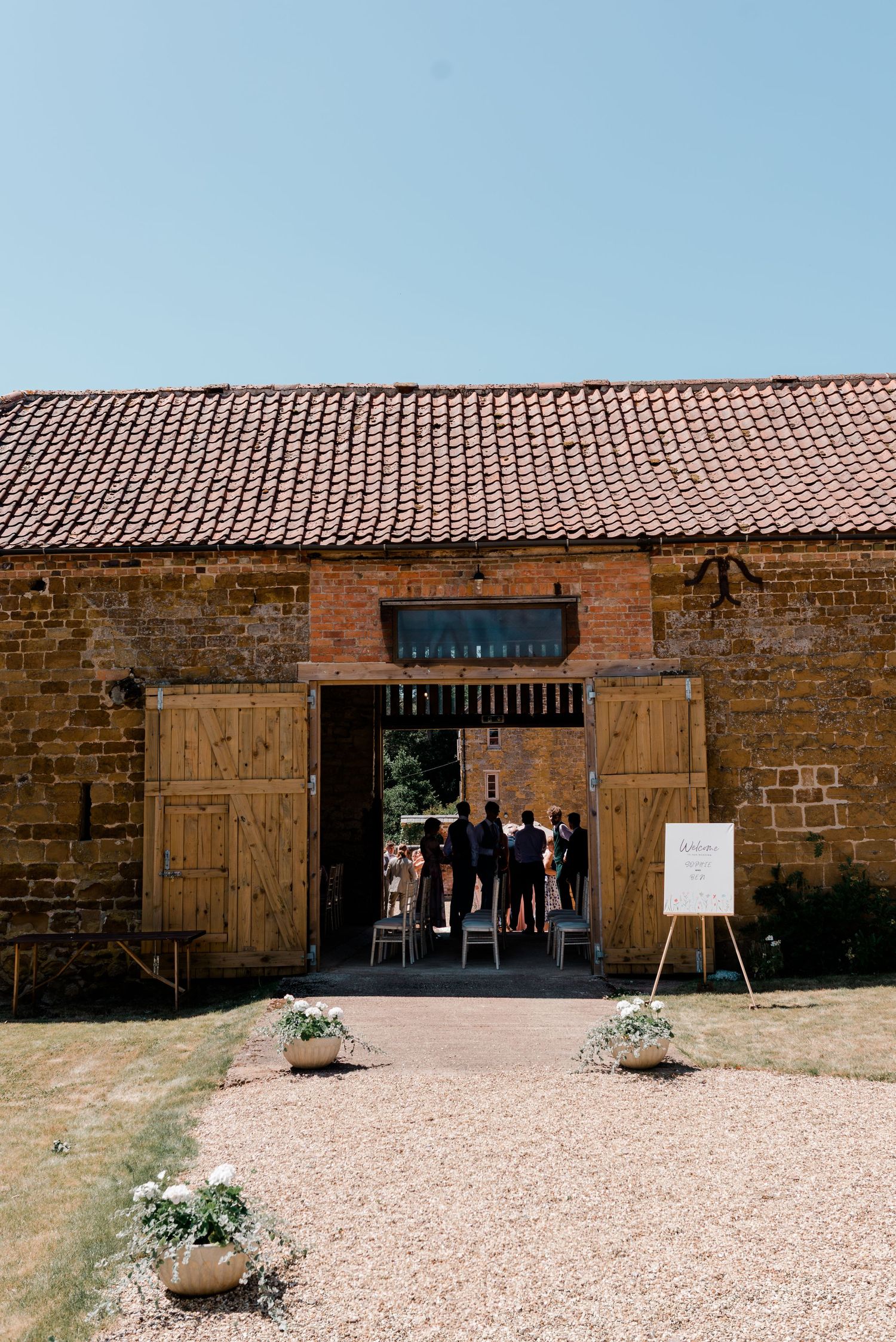 Rustic brick barn venue with terracotta roof tiles and wooden doors on a sunny day.