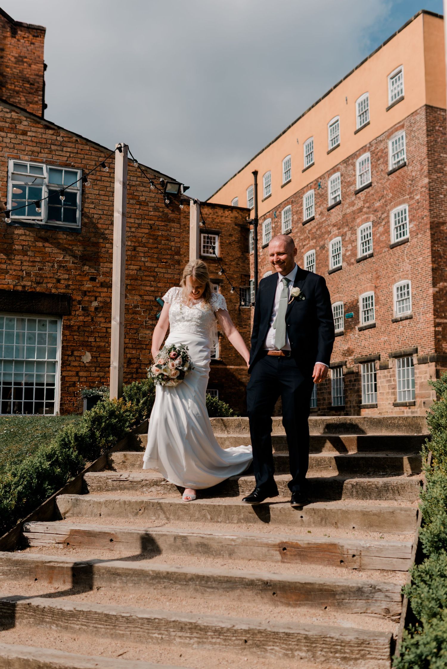 Bride and groom descending stone steps hand in hand outside a grand red brick mill venue.