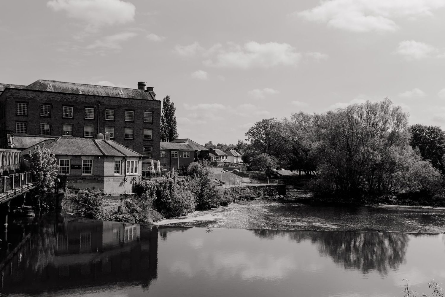 Black and white photo of a historic mill building beside a tranquil river surrounded by trees.