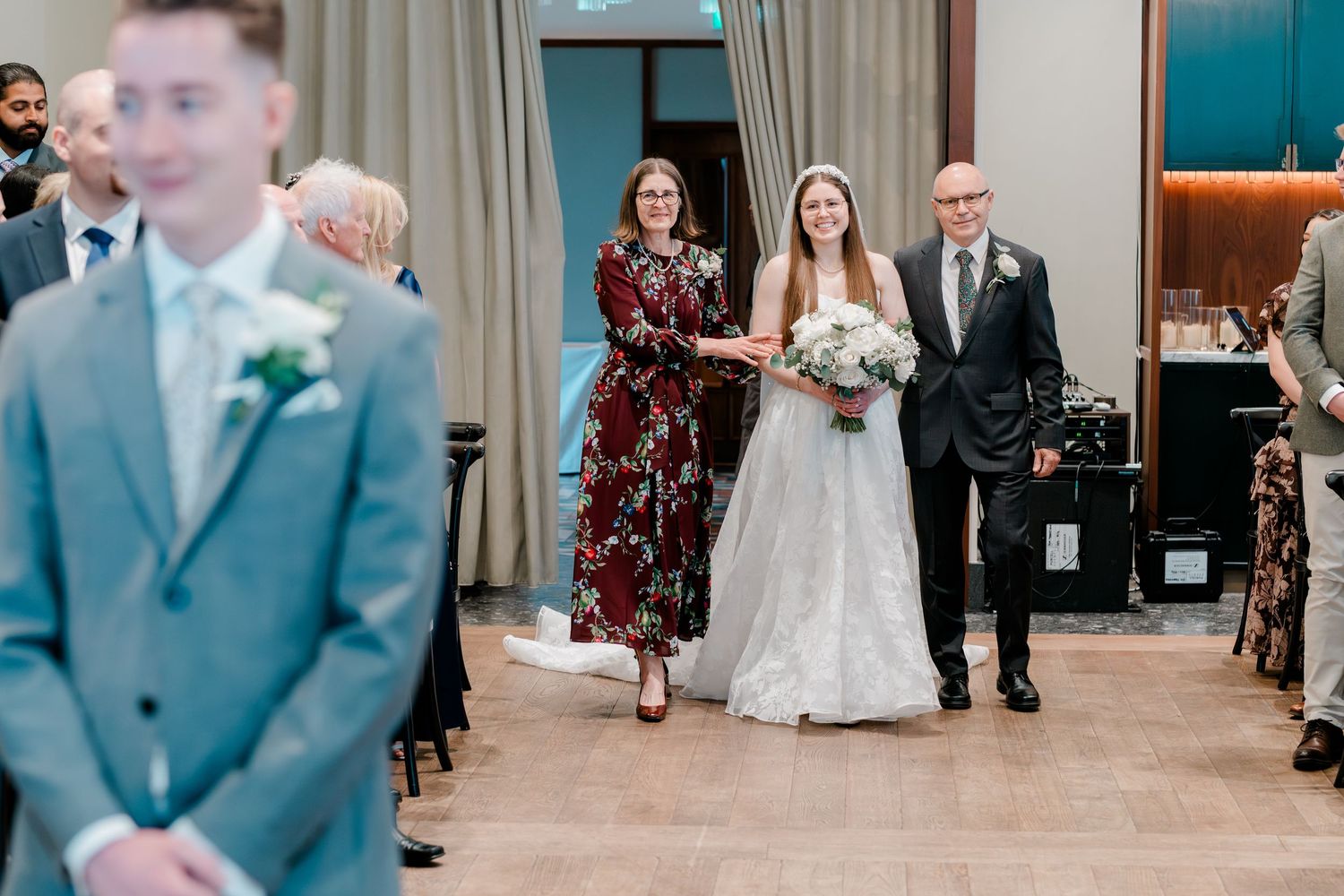 Bride walks down the aisle with parents during elegant indoor wedding ceremony.