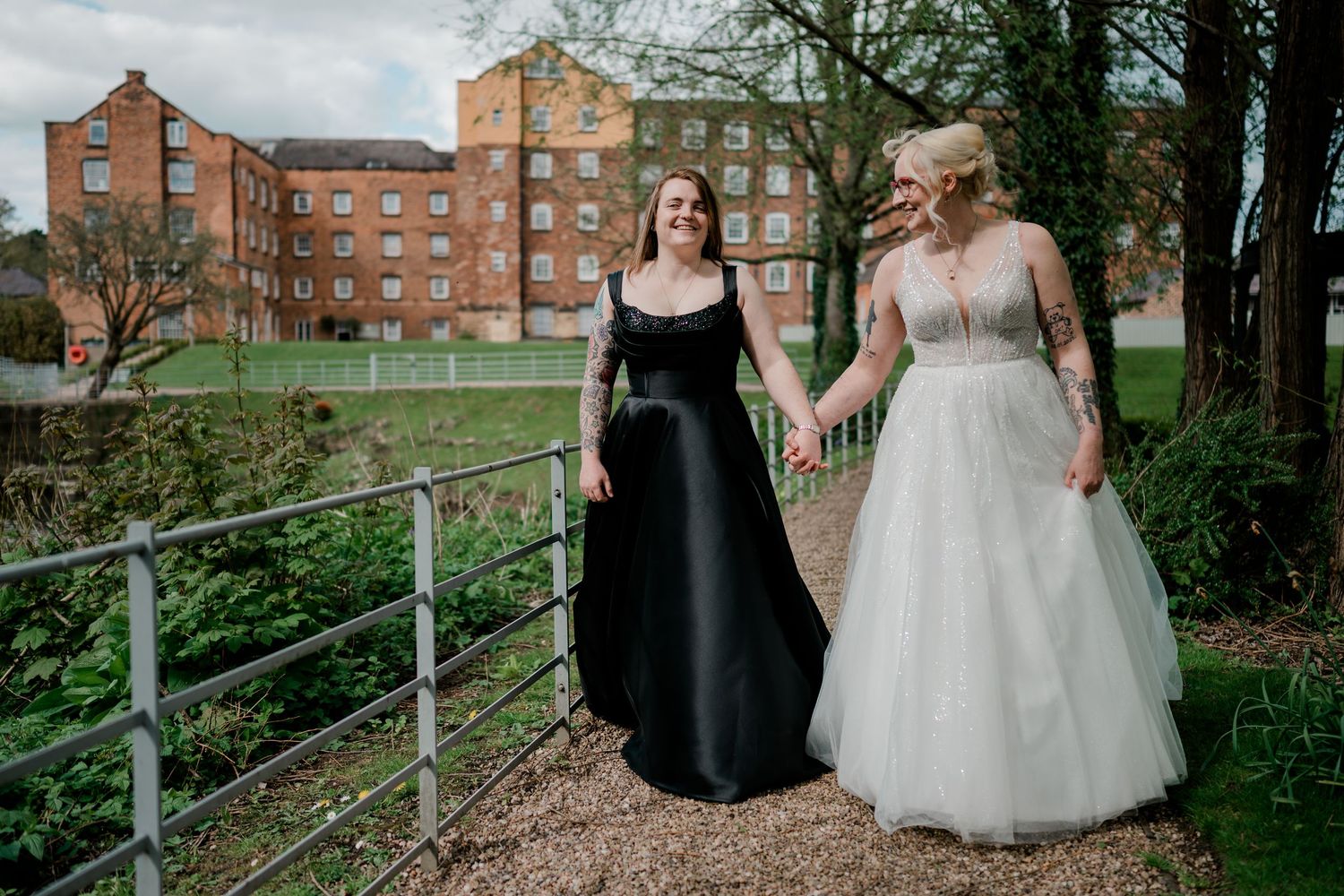 Two brides holding hands walking outdoors, one in white ballgown and one in black lace dress.