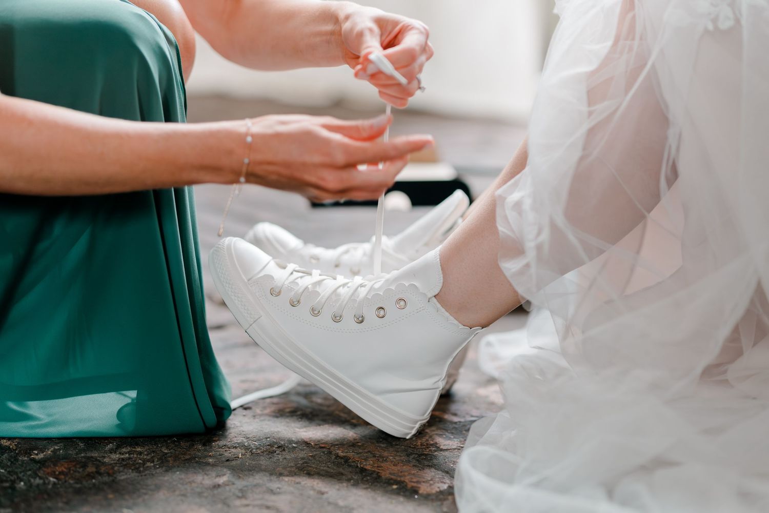 Bridesmaid in teal dress helps bride lace up white high-top Converse sneakers on wedding day.