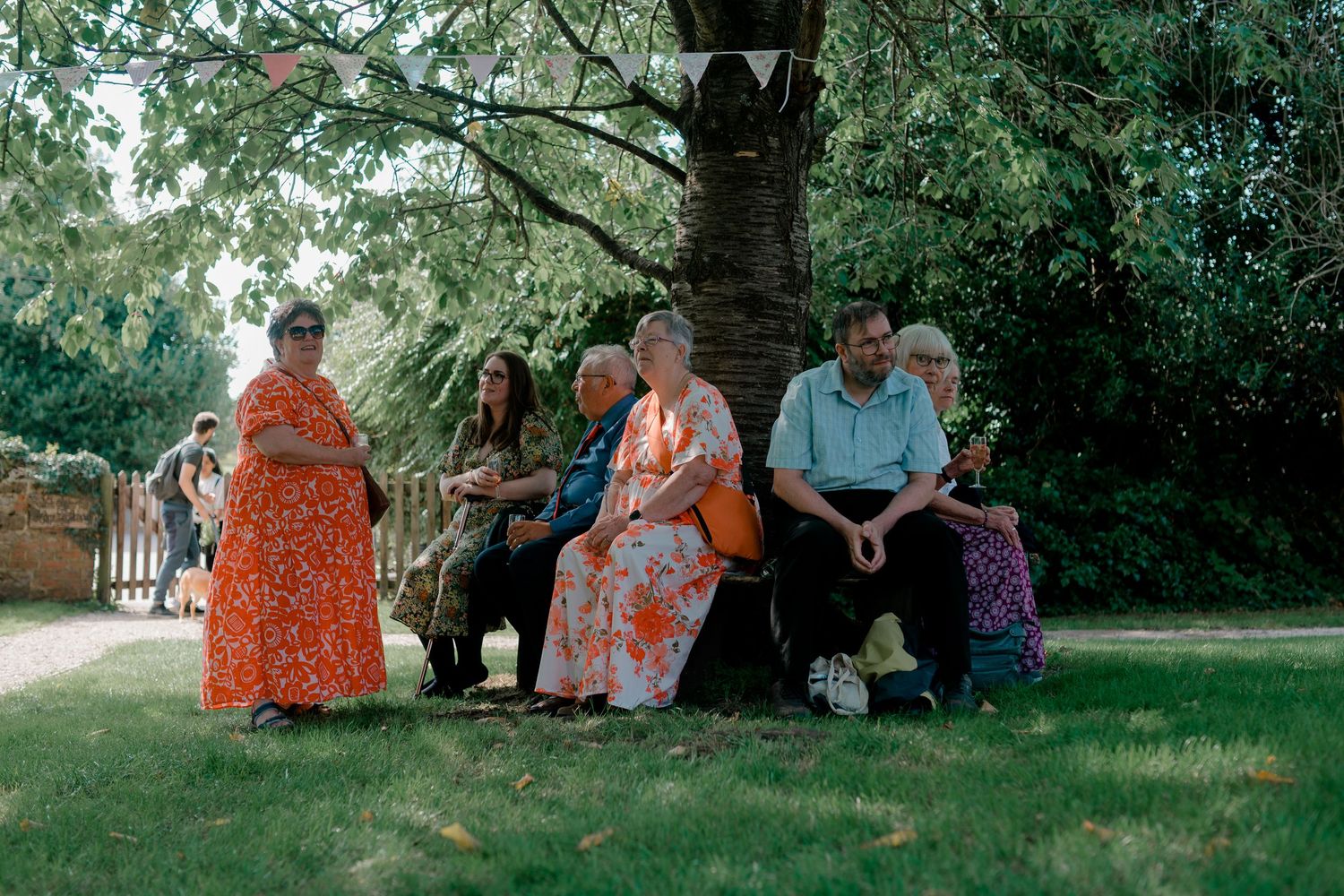 Group of wedding guests sitting together under large trees in garden setting.