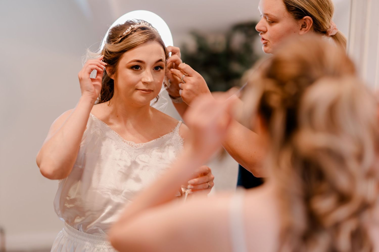 A bride puts on delicate earrings while getting ready in a soft, warm-lit room.