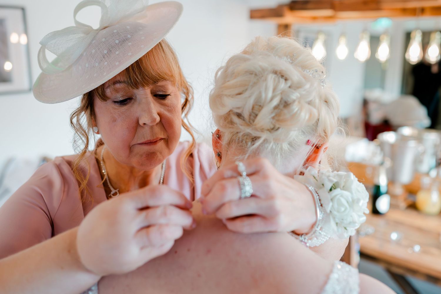 Woman in pink dress and fascinator fastening the back of a bride's lace wedding gown before the ceremony.