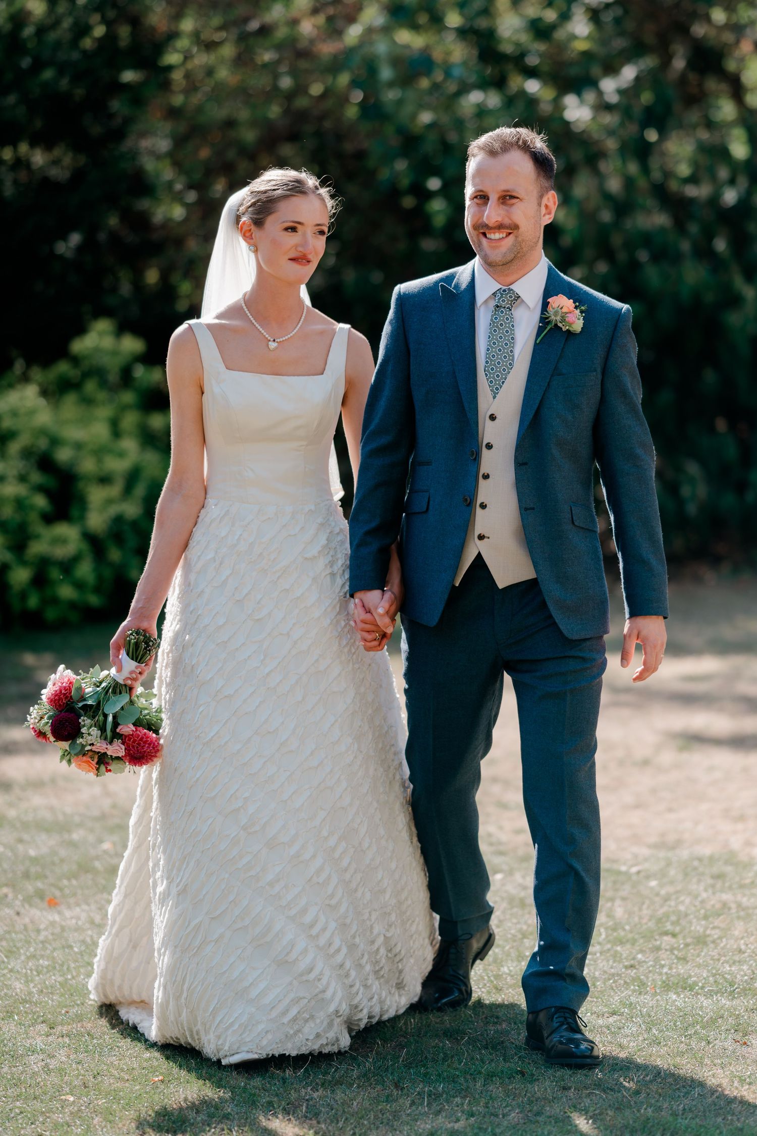 A bride in a white textured gown and groom in a navy suit walk hand in hand outdoors on their wedding day.