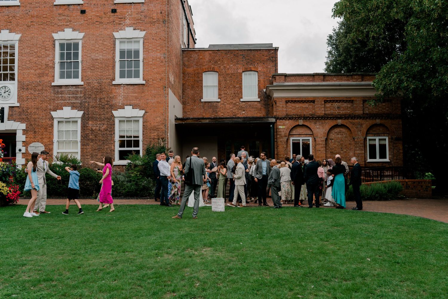 Wedding guests gather on a green lawn outside a historic red brick venue during an outdoor wedding reception.