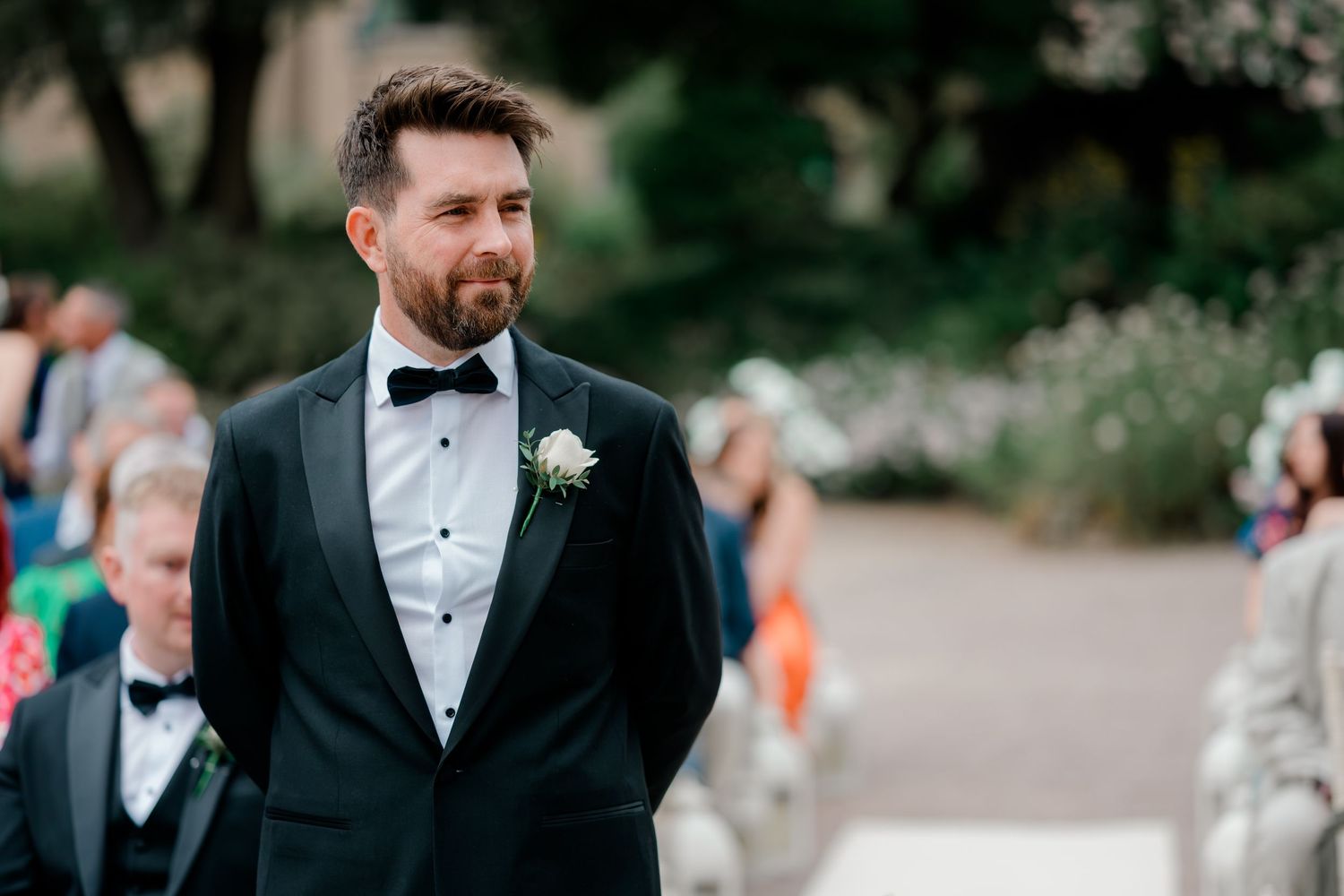 Groom in a black tuxedo with bow tie and white boutonniere stands at an outdoor wedding ceremony with guests seated behind him.