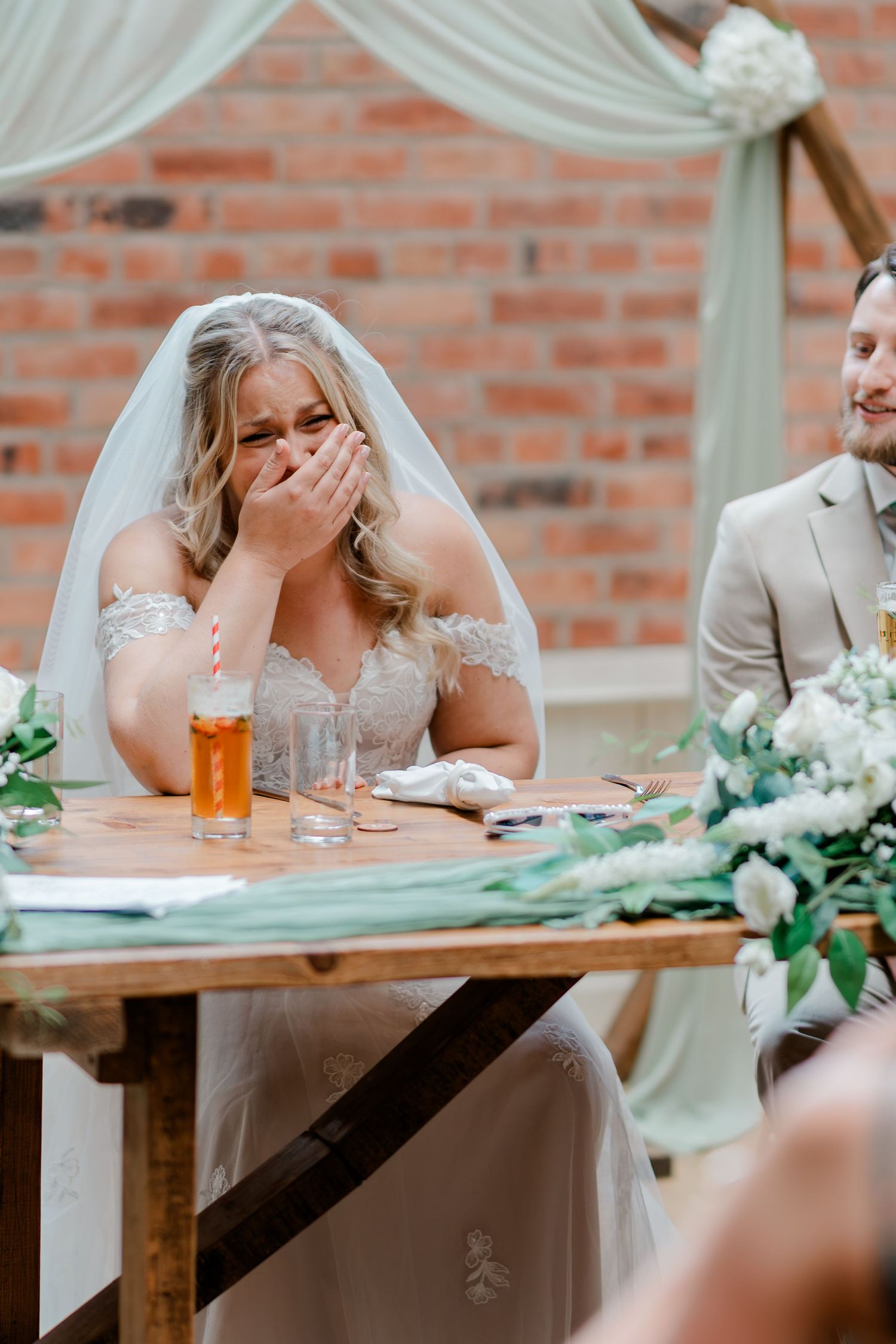 Bride laughs joyfully at sweetheart table during wedding reception speeches beside smiling groom.