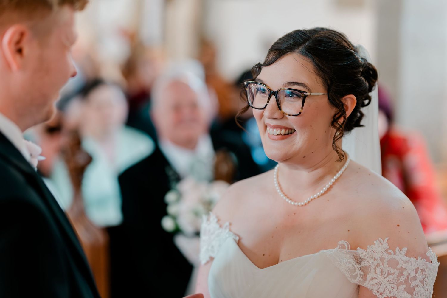 Bride in off-shoulder lace gown and glasses smiling at groom during wedding ceremony in church.