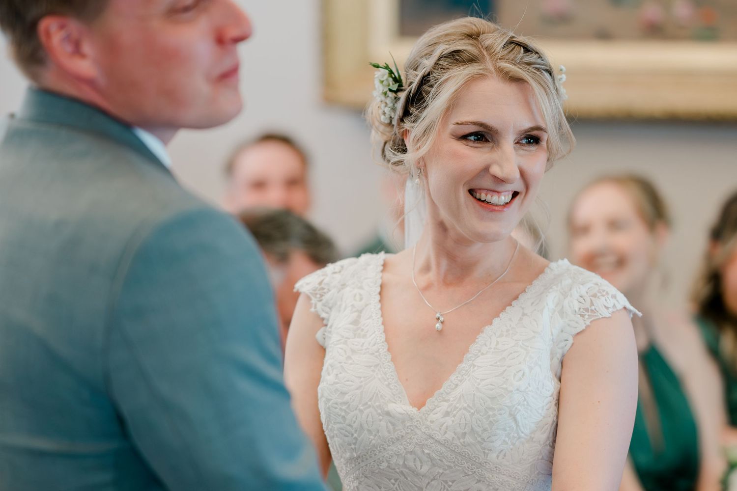 Radiant bride in lace wedding dress smiling joyfully during wedding ceremony with guests in background.