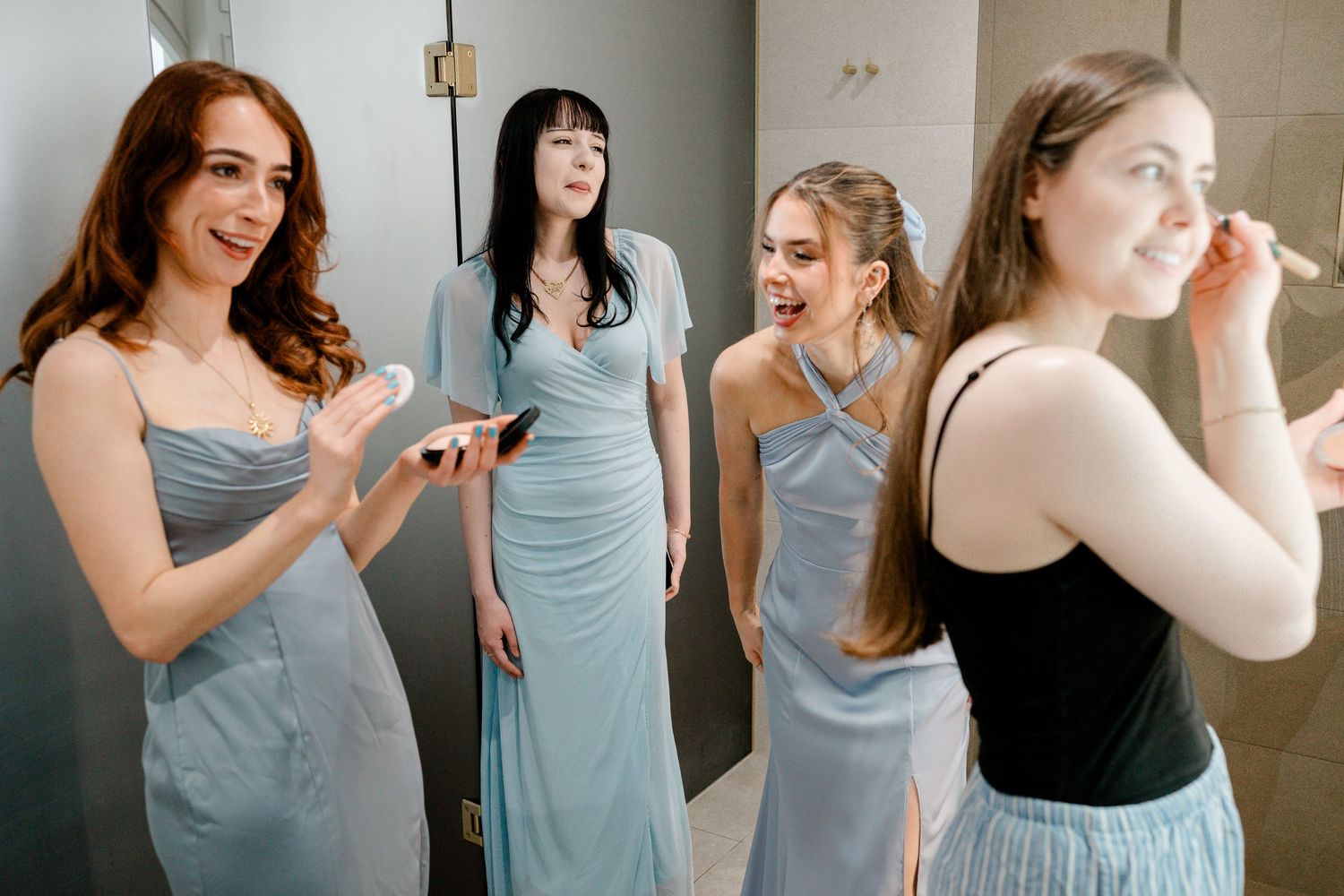 Four bridesmaids in light blue gowns laugh and apply makeup together while getting ready for a wedding.