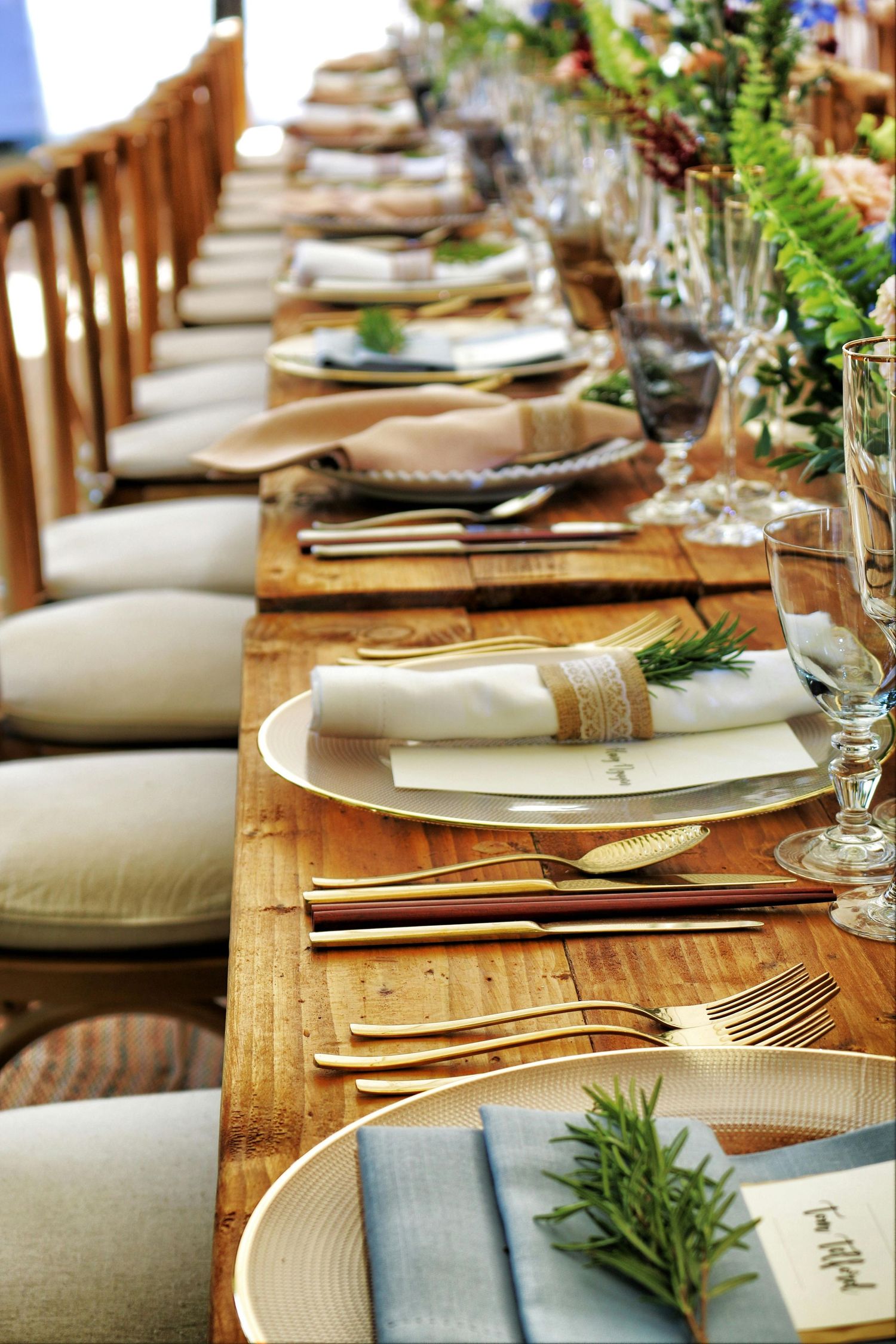 Elegant wedding table setting with wooden tables, gold cutlery, white plates and fresh greenery decorations.