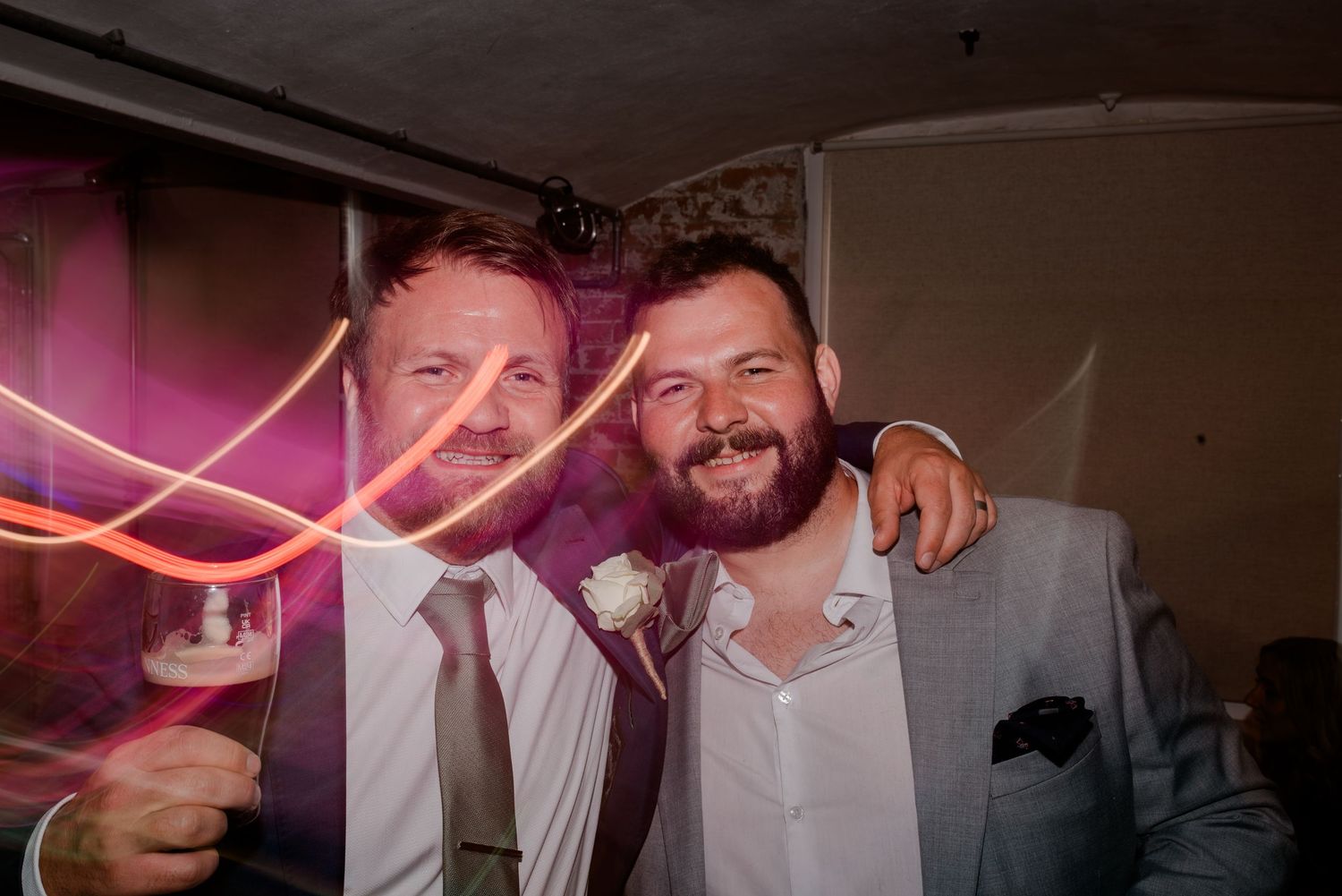 Two men celebrate at a party with neon pink lighting and festive atmosphere.
