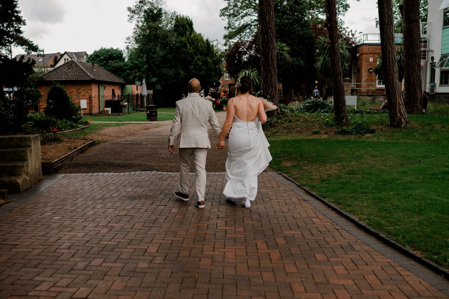 Couple walking along a brick pathway surrounded by green trees and buildings at dusk.