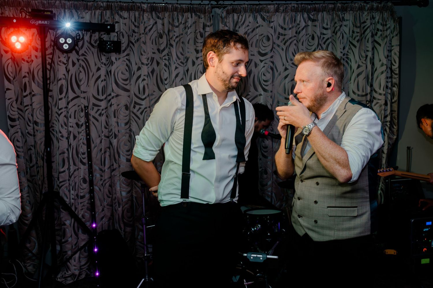 A person in a gray vest and white shirt dances energetically on a dark dance floor with patterned wallpaper.