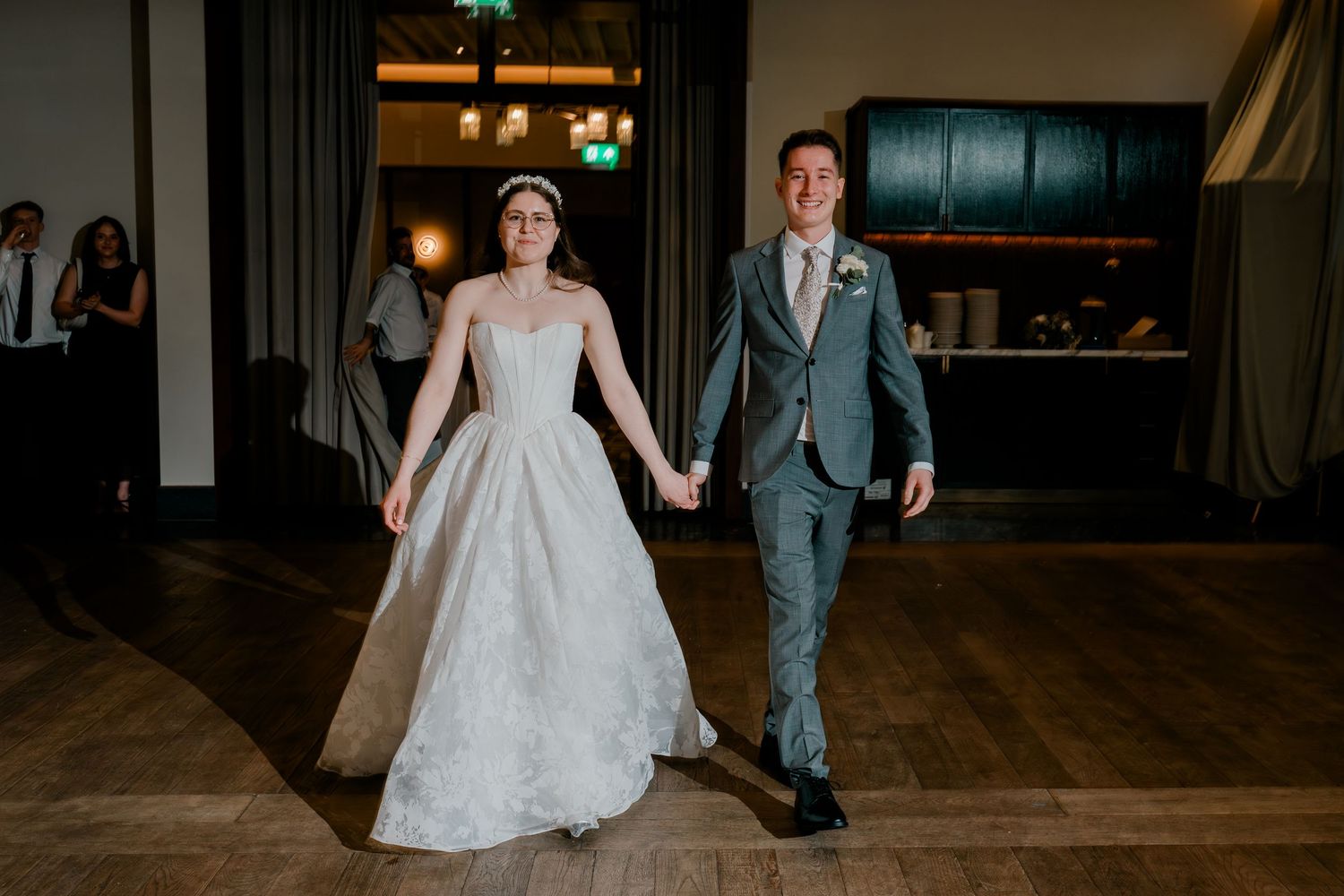 Bride in white gown and groom in gray suit hold hands entering wedding reception on wooden dance floor.
