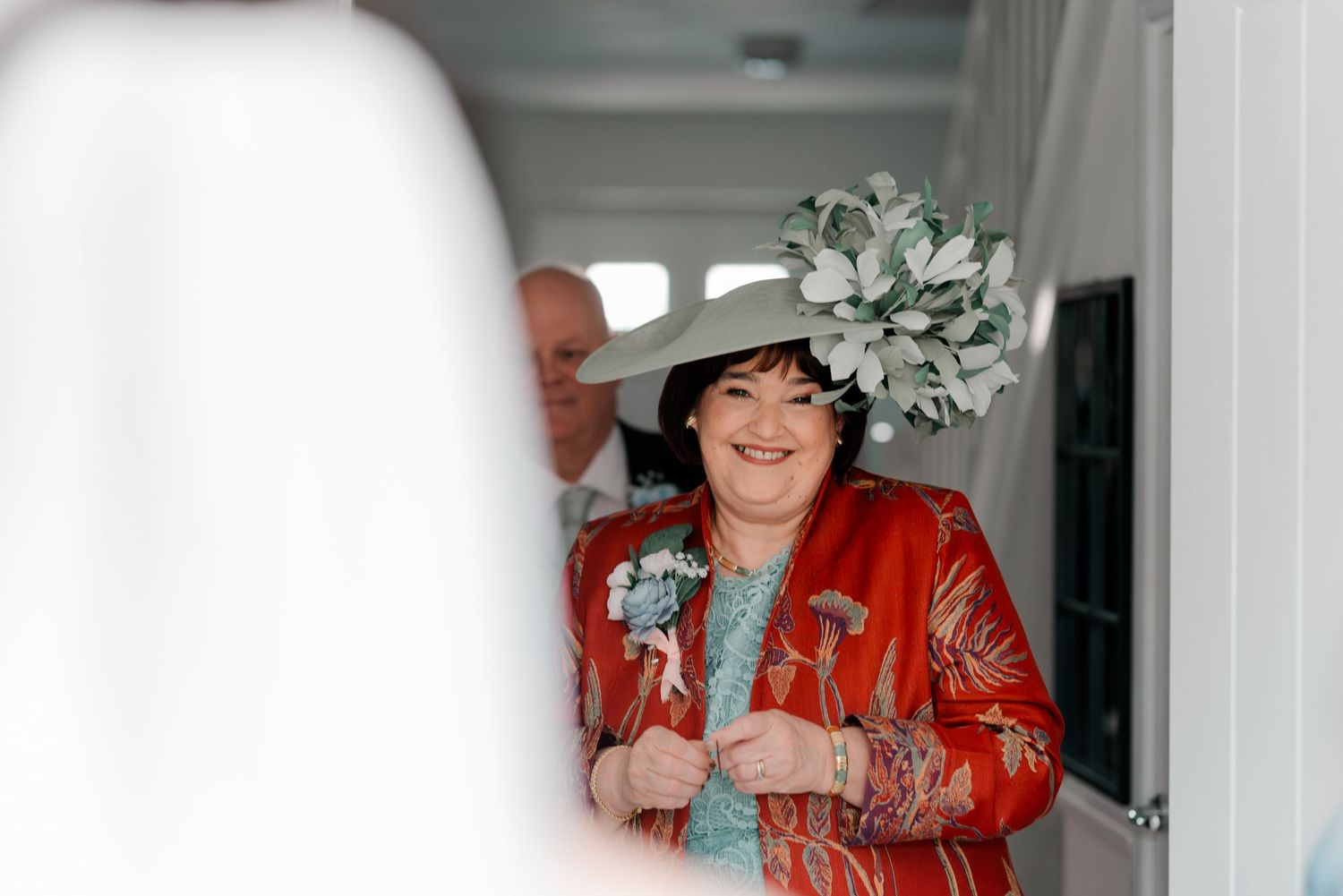 A smiling woman in a red floral jacket and elegant white fascinator hat at a wedding celebration.