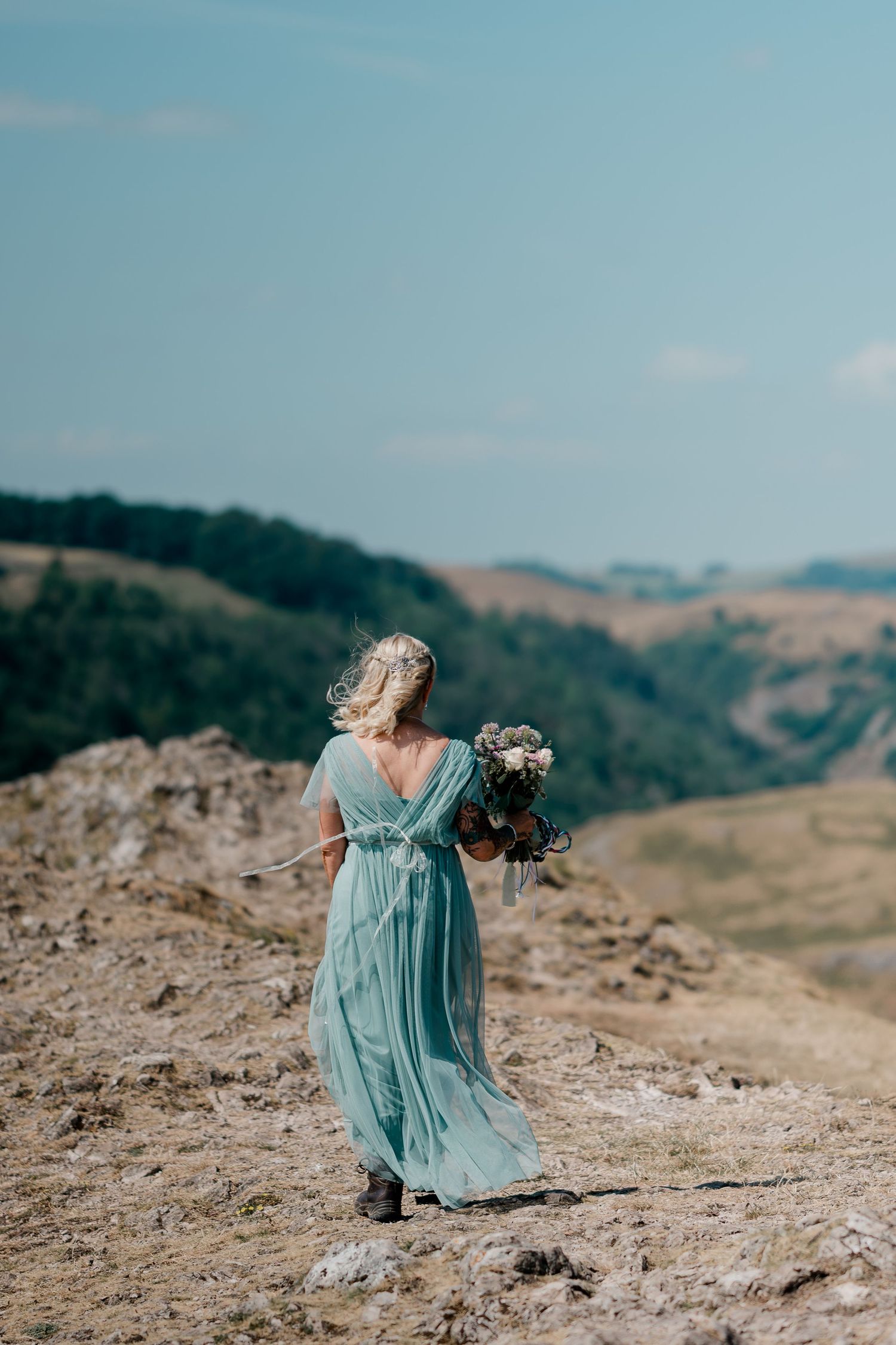 Bride in flowing teal dress holds bouquet while standing on a scenic hilltop.