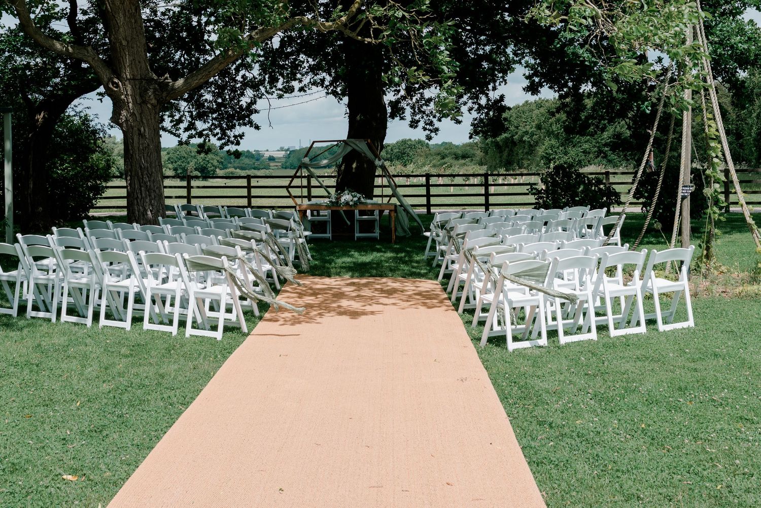 Outdoor wedding ceremony setup with white folding chairs, tan aisle runner, and wooden arch on lush green lawn.
