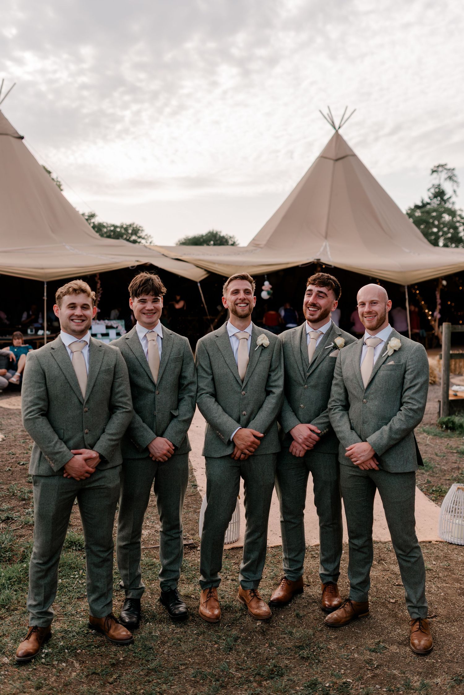 Groomsmen in matching sage green suits pose together in front of outdoor tipi-style wedding tents.