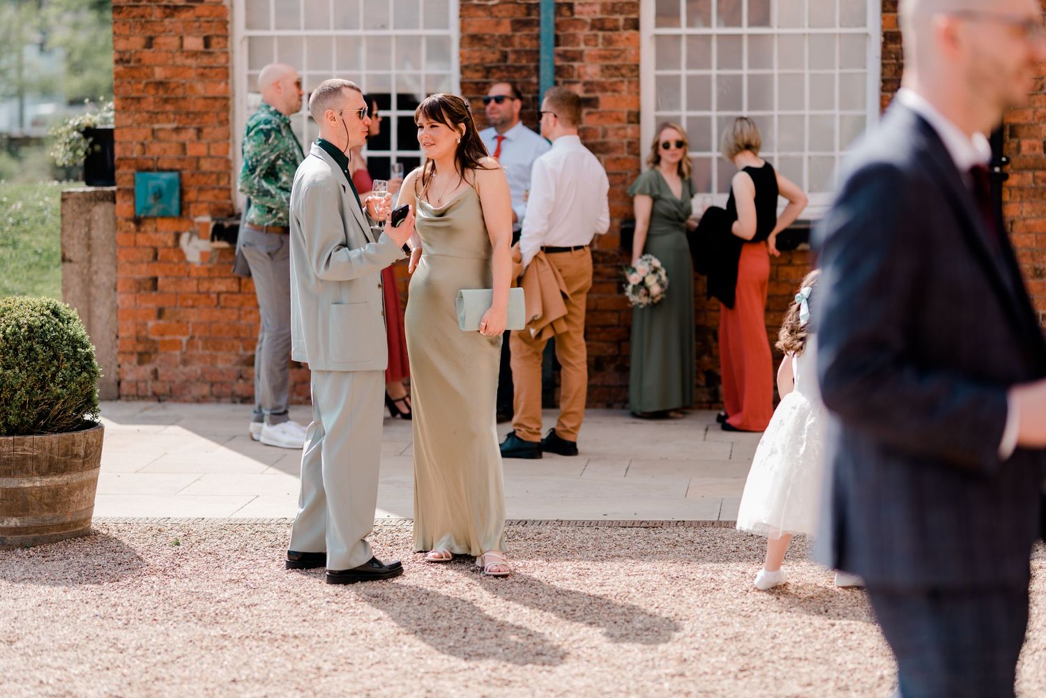 Wedding attendees socializing outside historic brick venue with white windows and gravel courtyard.