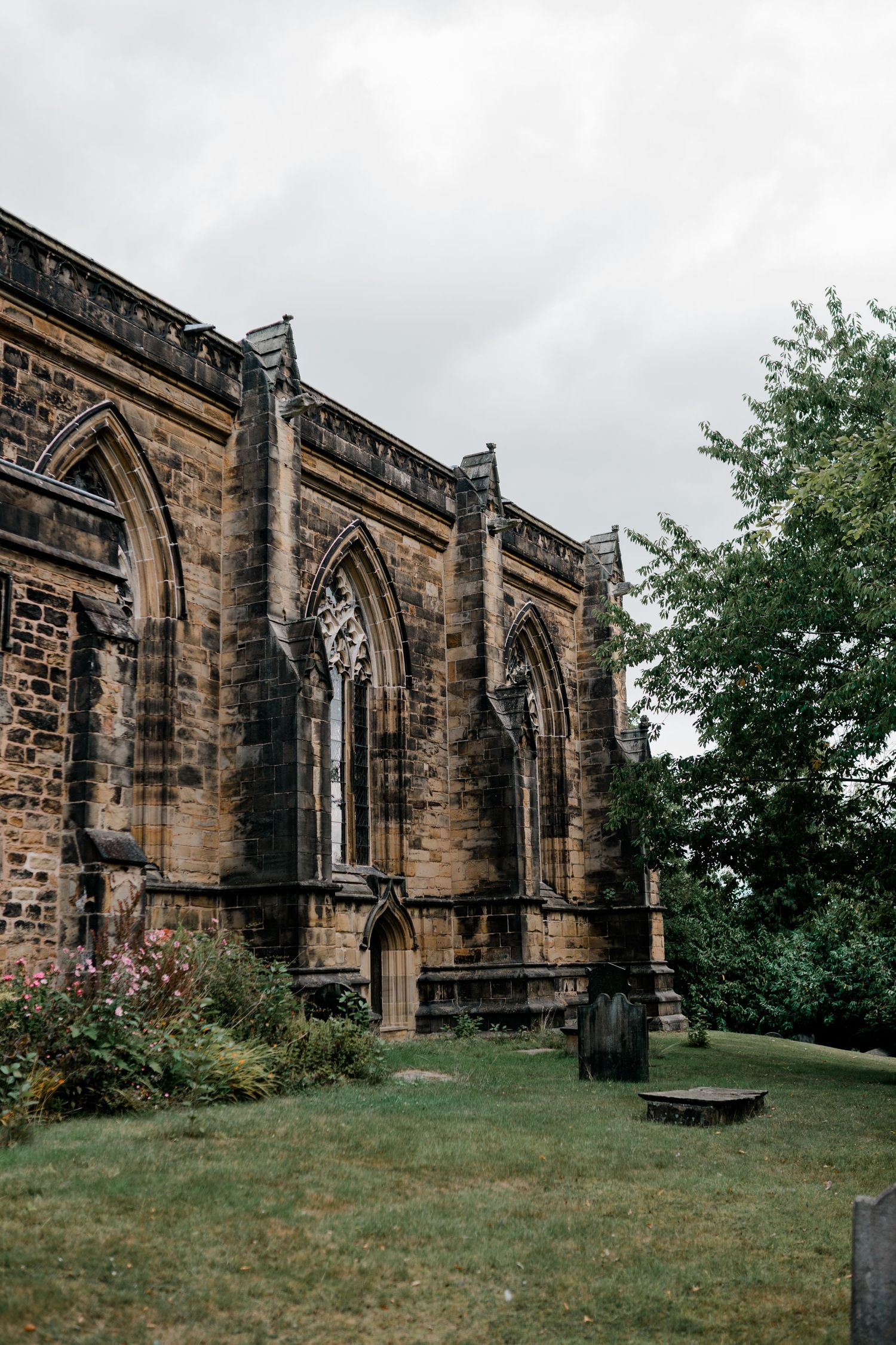 Gothic stone church exterior with arched windows and buttresses set against cloudy sky and green lawn.