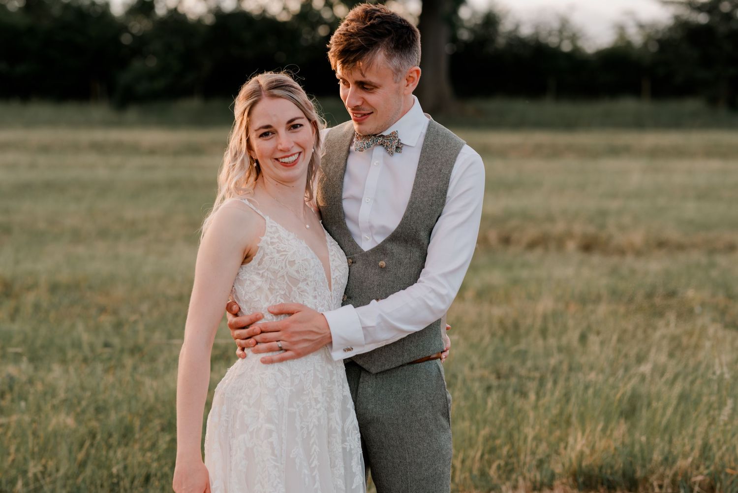 Smiling bride and groom posing together in a sunlit meadow, groom wearing a grey waistcoat and bow tie.