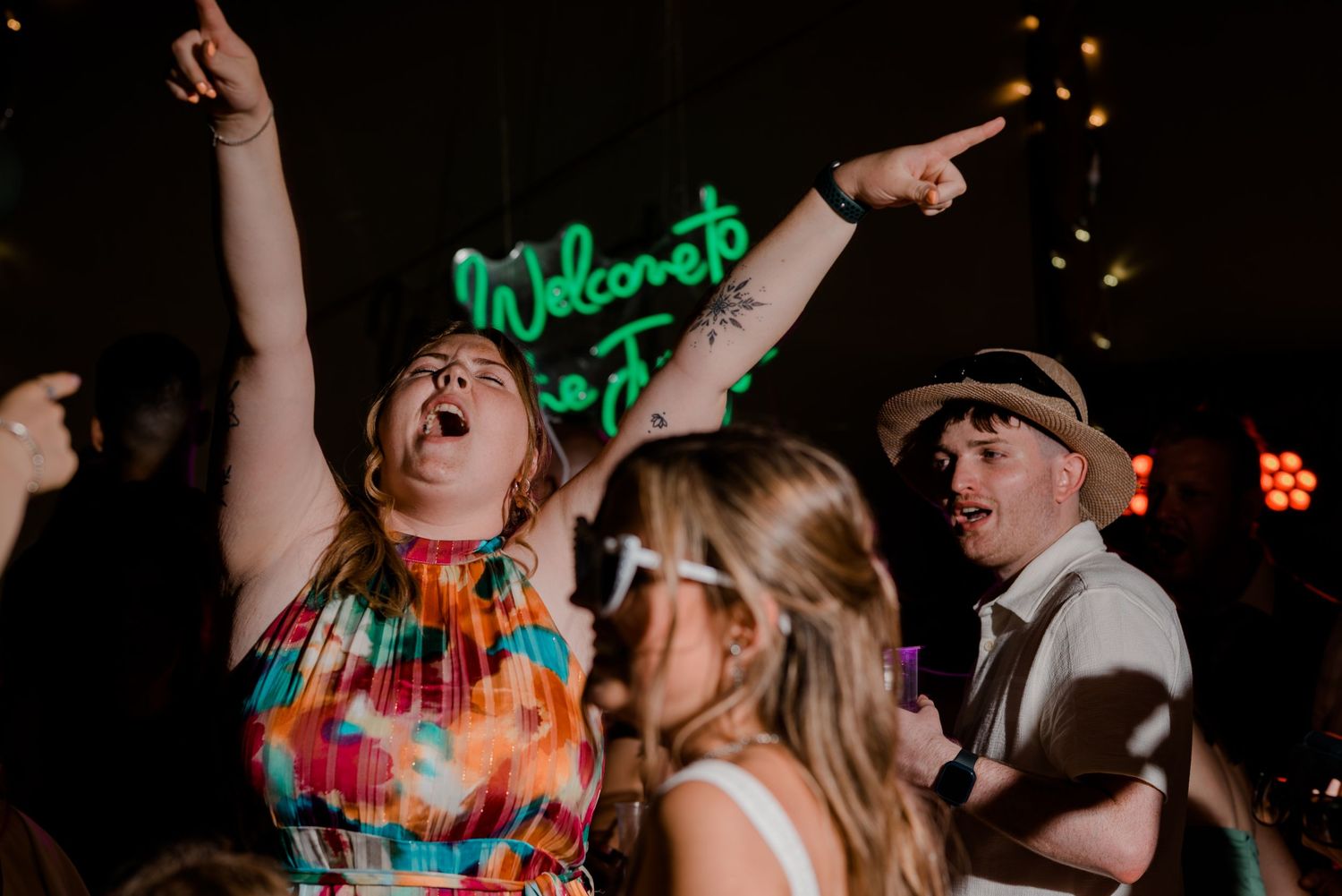 Energetic wedding guests dancing under a Welcome to the Jungle neon sign at a lively reception.